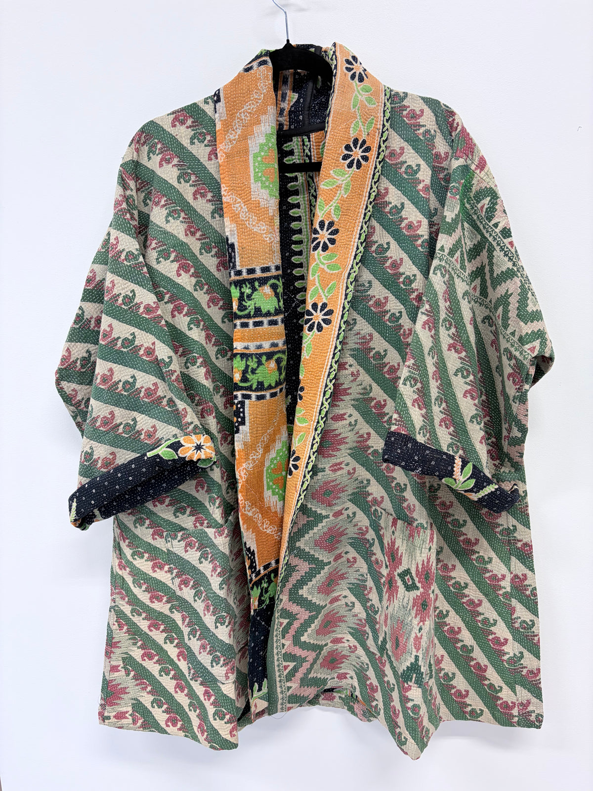 A patterned open-front kimono-style cardigan or robe hanging against a plain white background. The garment features green diagonal stripes, pink floral motifs, and a wide orange-gold patterned lapel with black and green accents.