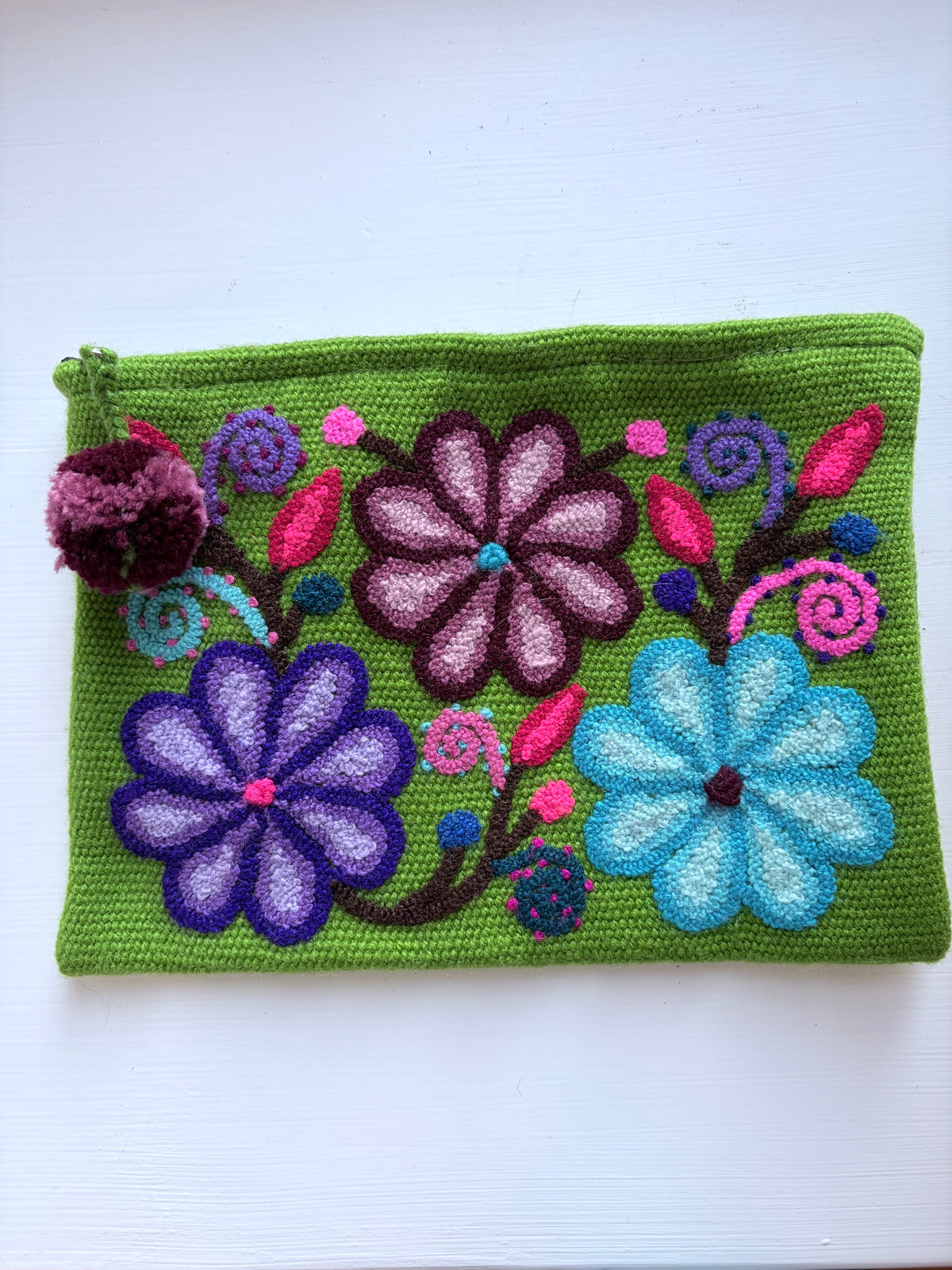 A green knitted pouch featuring colorful hand-embroidered flowers in purple, blue, and pink, with a small matching pom-pom charm on the left side, set against a plain white background.