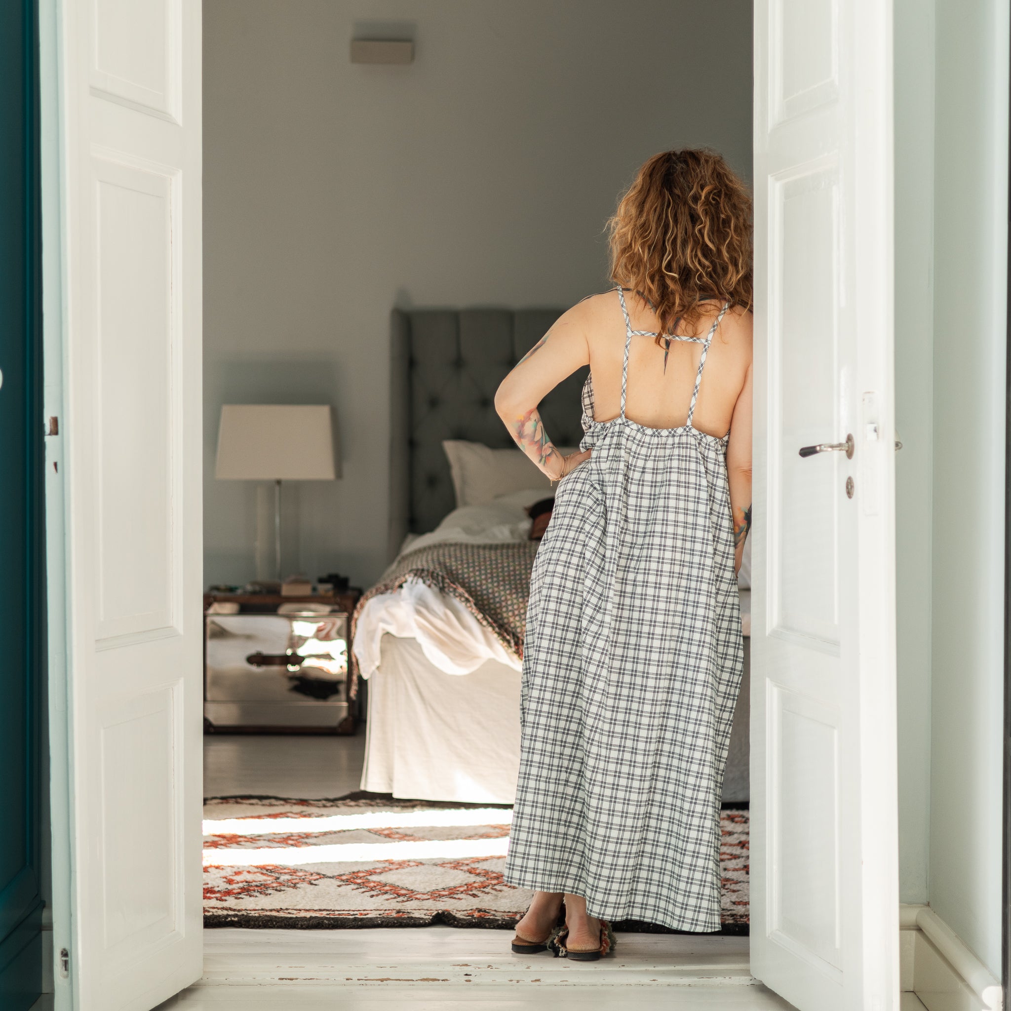 Woman in a checkered dress standing in a doorway looking into a bedroom.