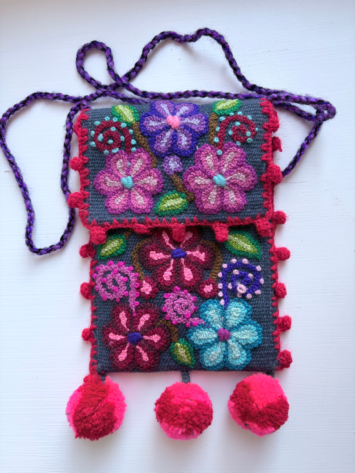 A handmade crocheted shoulder bag with a dark gray base, featuring colorful embroidered floral patterns in pink, purple, blue, and green. The bag has a purple braided strap and is decorated with bright pink pom-poms along the edges and three large pom-poms hanging from the bottom.