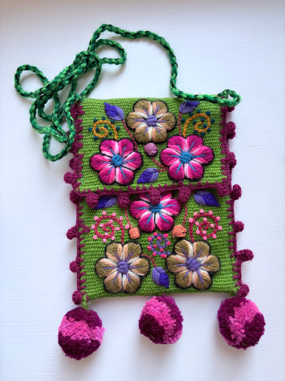 A green crocheted shoulder bag with colorful floral embroidery, pink pom-poms, and a green braided strap, laid flat on a white background.