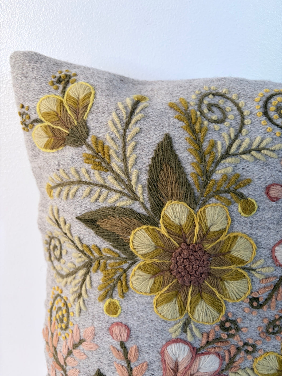 Close-up of a square decorative throw pillow in light gray fabric with intricate floral embroidery in yellow, olive green, brown, pink, and cream, set against a plain white background.