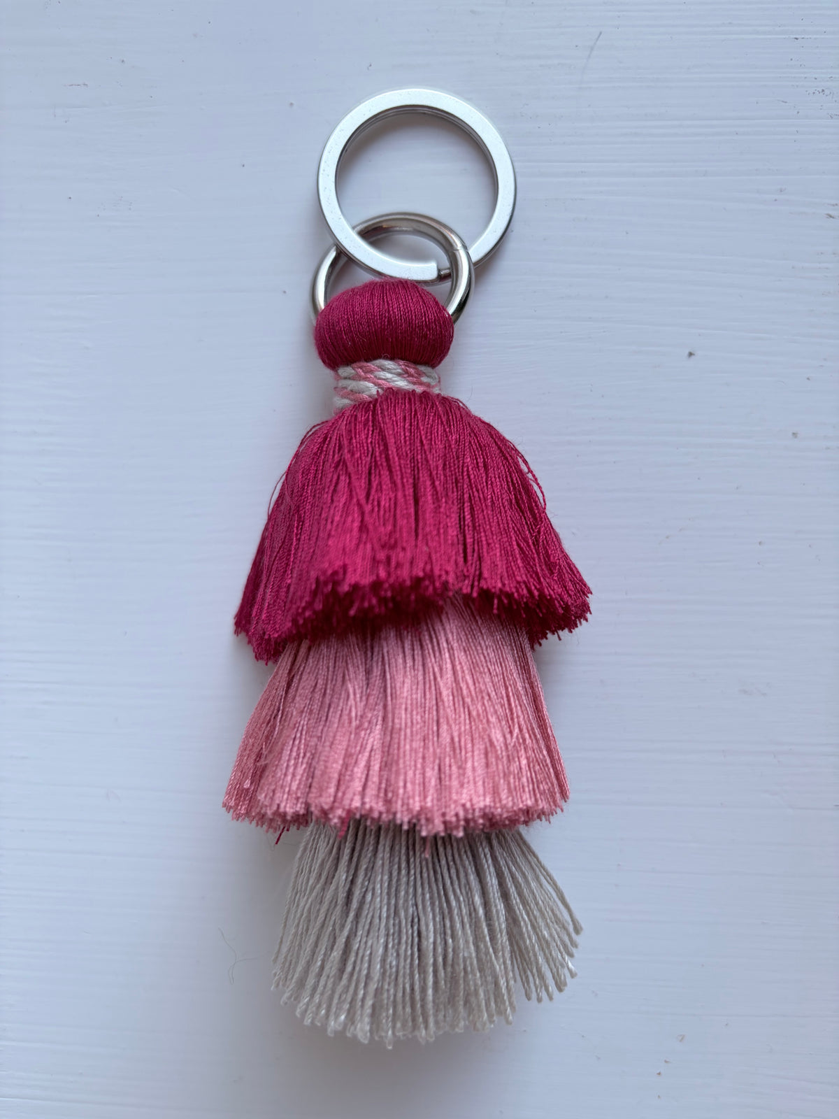 A decorative tassel keychain with three stacked tiers of tassels in dark magenta, light pink, and off-white, attached to a silver split ring on a light gray background.