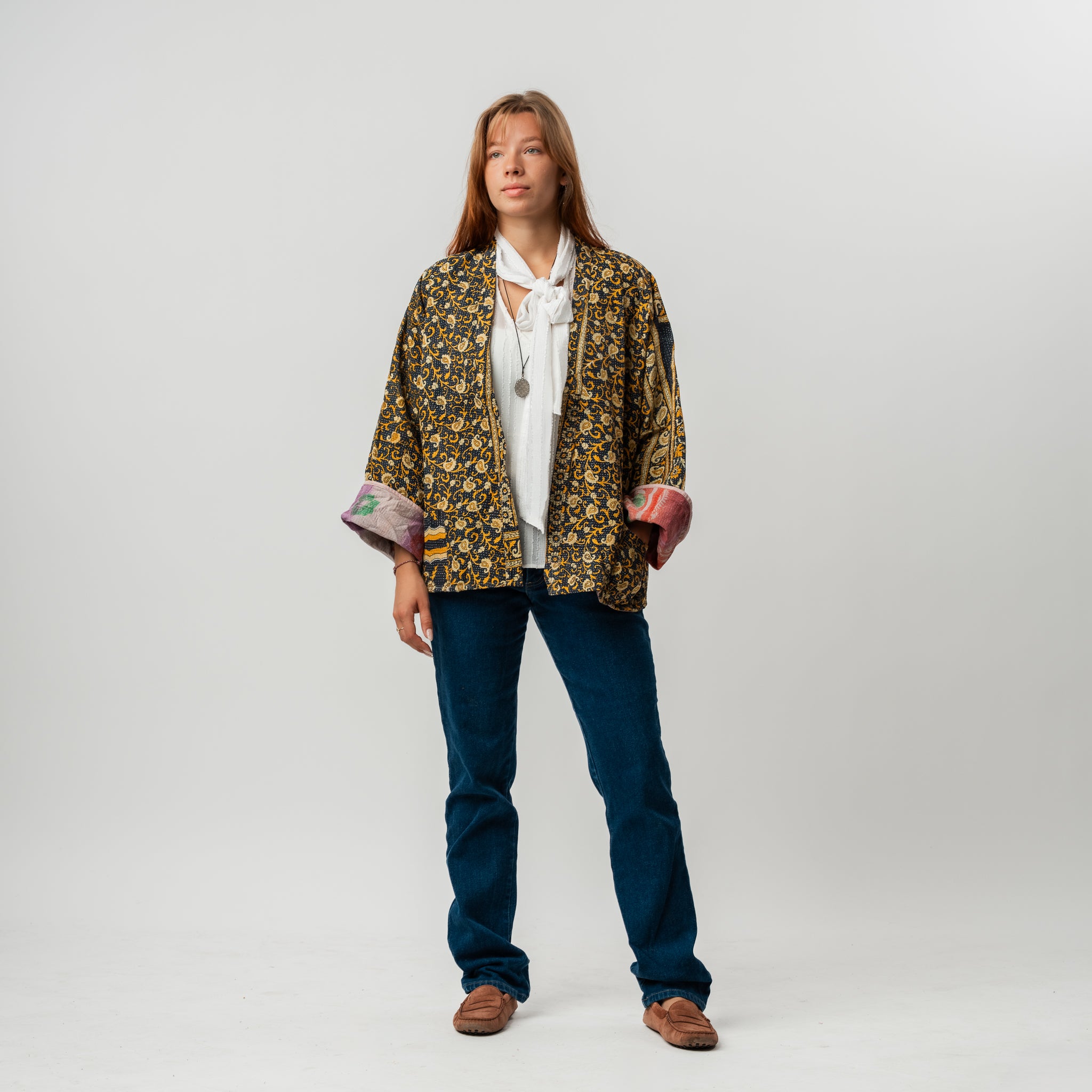 Woman wearing a patterned jacket, white shirt, blue jeans, and brown shoes on a white background