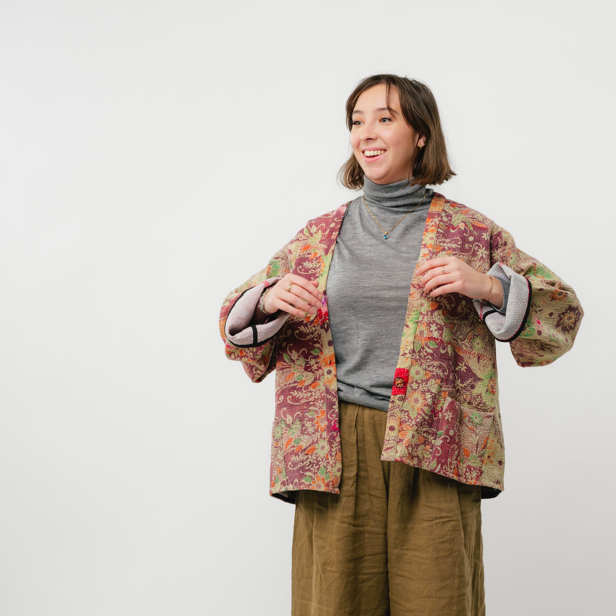 Woman wearing a colorful floral jacket over a gray turtleneck and brown pants on a white background