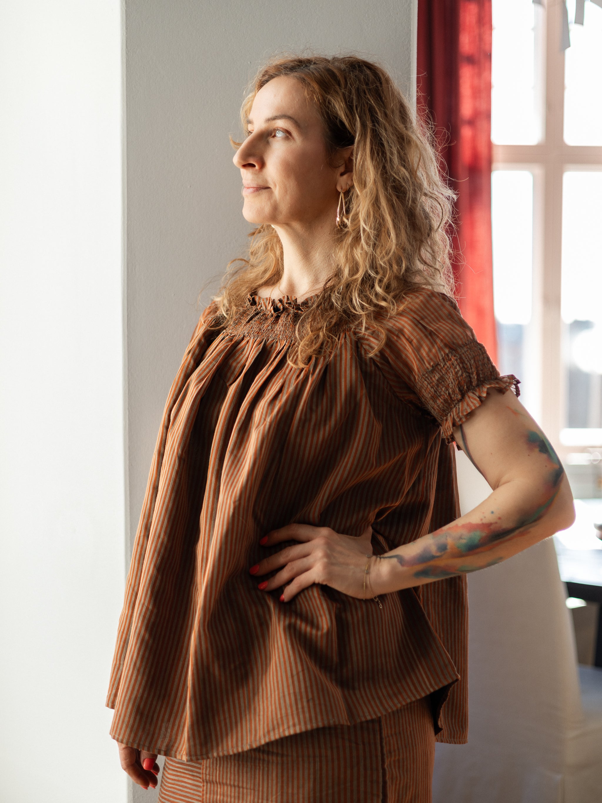 Woman wearing a brown pleated top and skirt standing indoors with a window in the background.