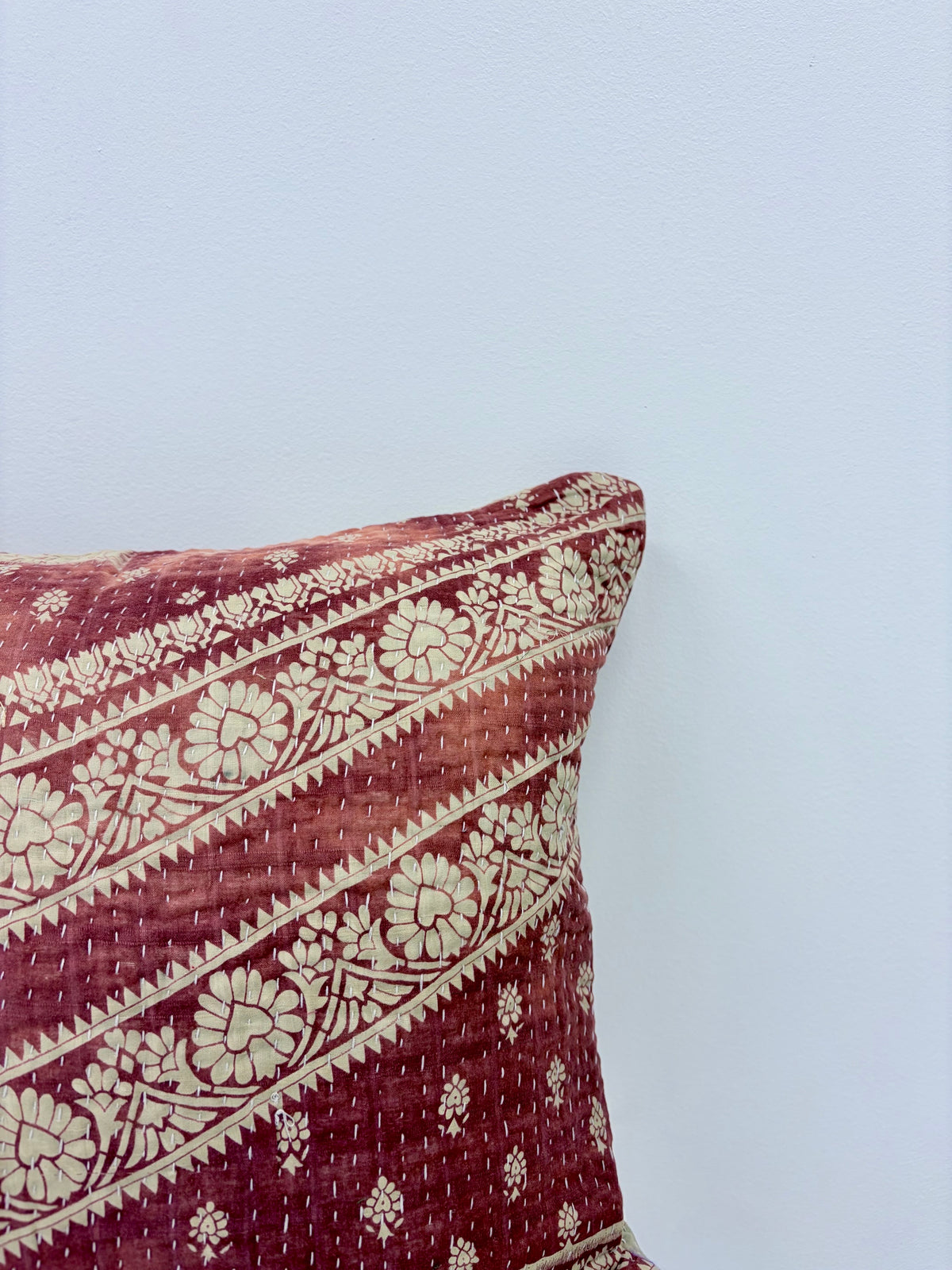 A close-up of a square throw pillow with a deep red and beige floral kantha quilt pattern, shown against a plain light blue wall.