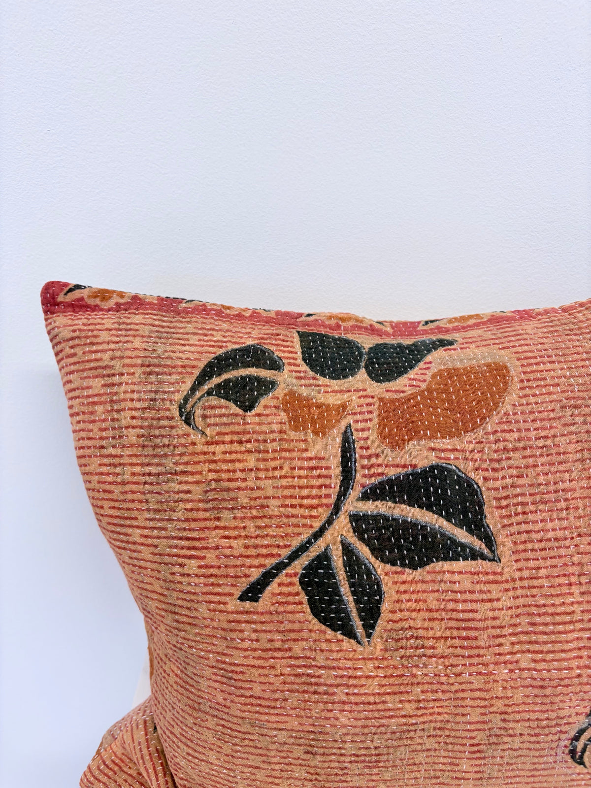A close-up of a textured throw pillow in a warm terracotta orange fabric, featuring a hand-embroidered floral motif with black and orange leaves.