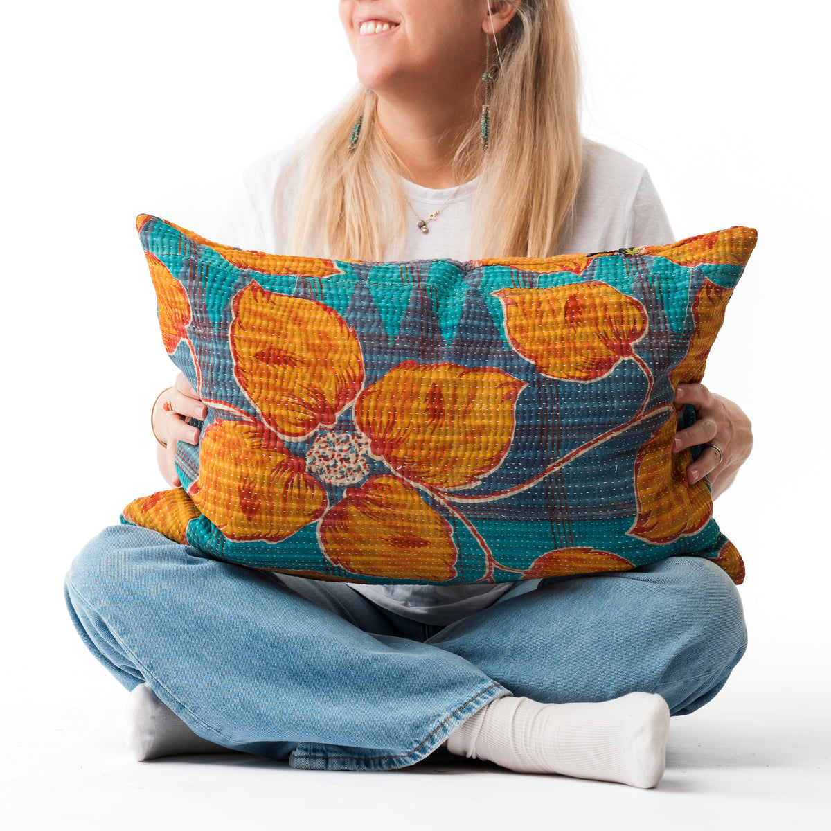 A woman sitting cross-legged on a white studio floor holds a square throw pillow with a vibrant orange and teal floral pattern.