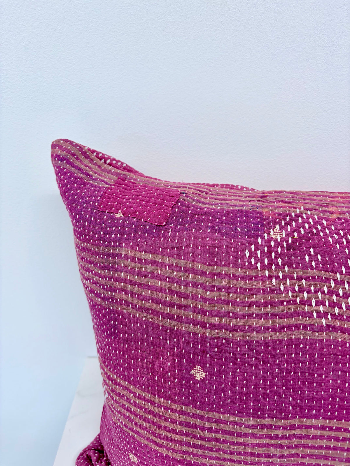 A close-up of a textured magenta and beige woven throw pillow, showing its handcrafted kantha quilt style with horizontal stitched stripes and small diamond motifs, set against a light blue wall.