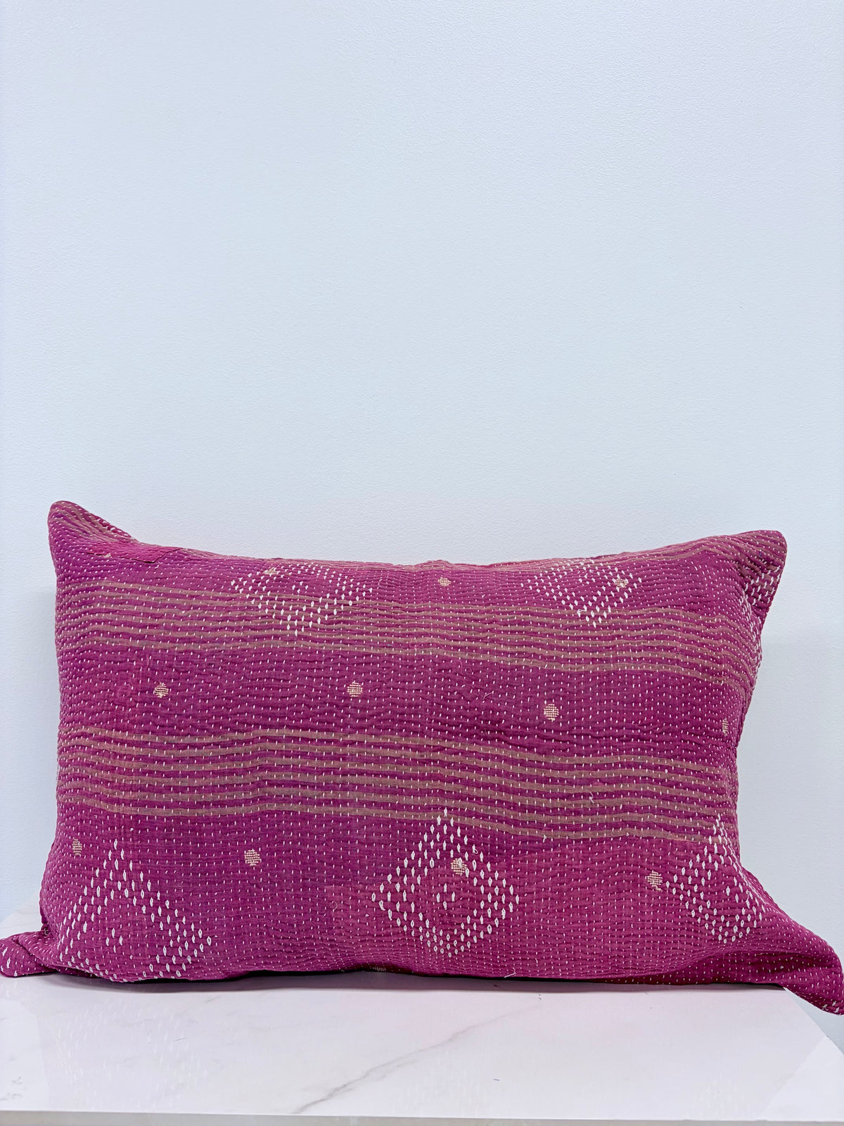 A rectangular woven throw pillow in a vibrant magenta pink with horizontal stripes and white geometric dot patterns, resting on a white marble surface against a light gray wall.