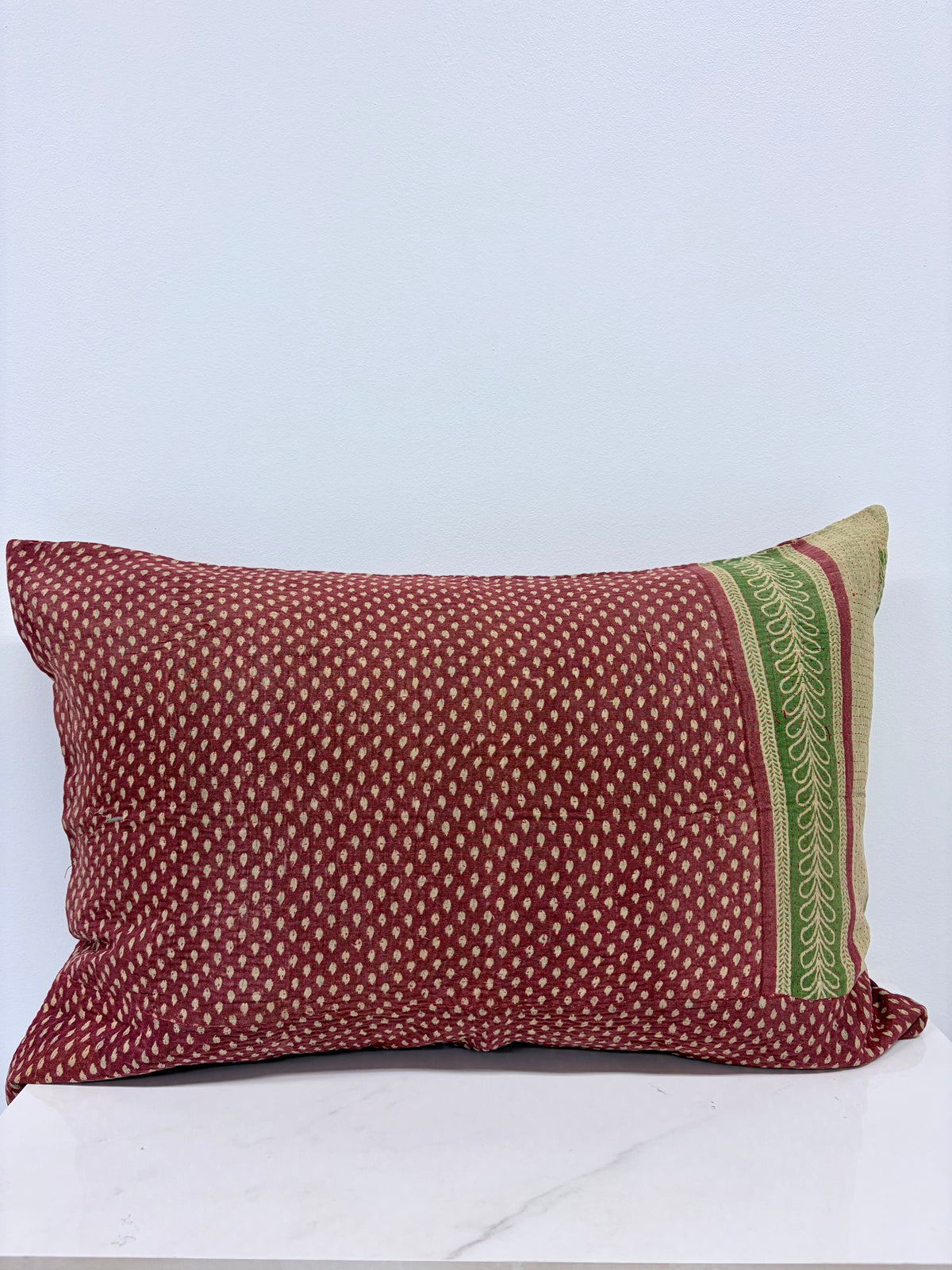 A rectangular throw pillow in rust red with a small white geometric pattern and a green and beige decorative border on one side, resting on a white marble surface against a light gray wall.