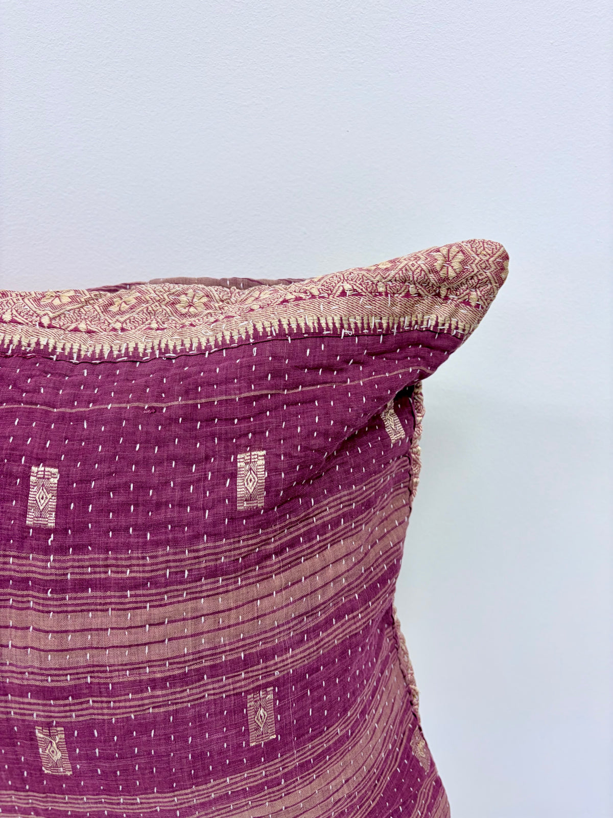 A close-up of a purple and beige kantha quilted throw pillow against a plain light blue-gray wall.