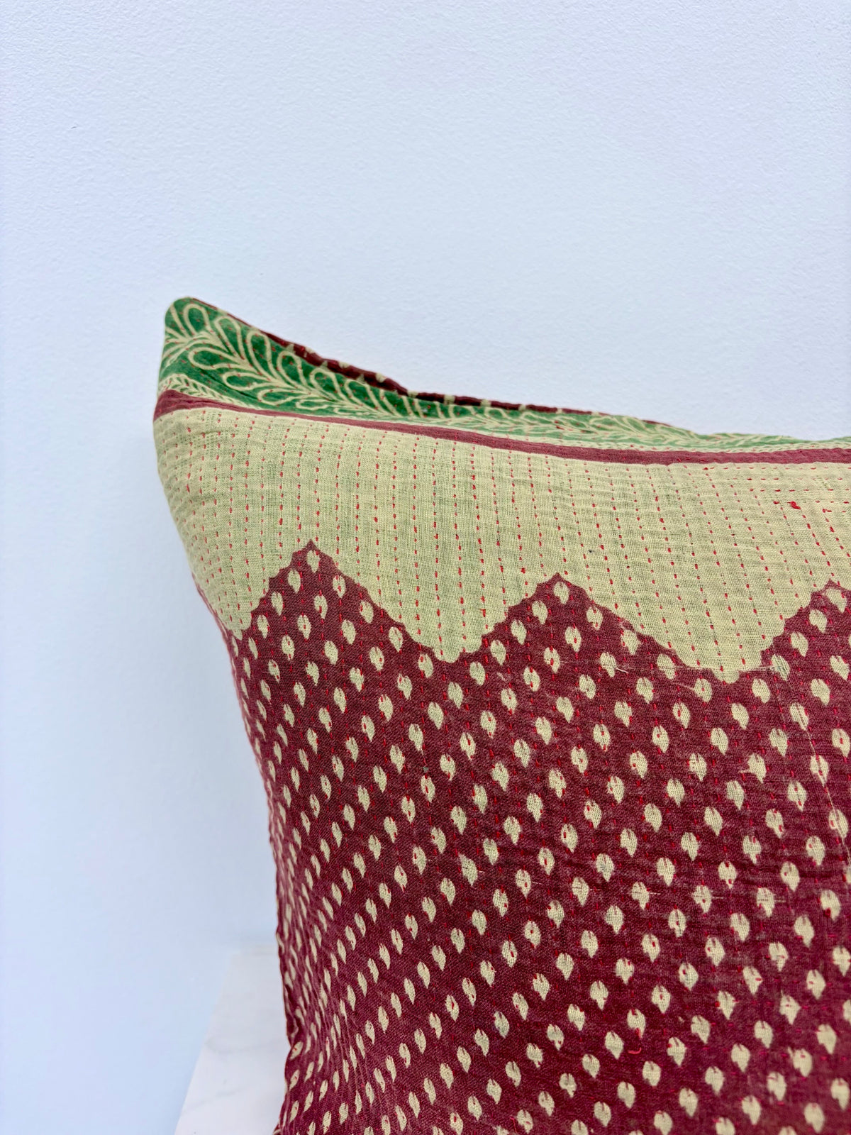 A close-up of a decorative throw pillow featuring a maroon and beige woven pattern with a green patterned border, set against a plain light blue wall.