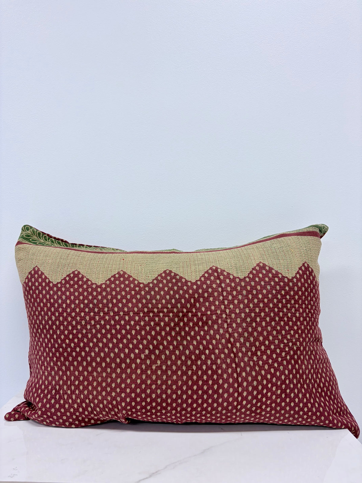 A rectangular throw pillow with a deep red body featuring a small white geometric pattern, a beige zigzag border, and a green patterned edge, displayed on a white marble surface against a light gray background.