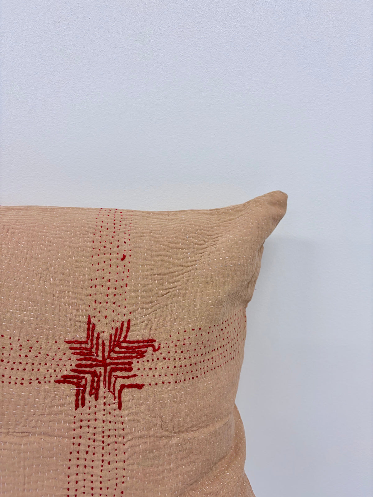 A close-up of a beige textured throw pillow with red embroidered geometric patterns, set against a plain white wall.