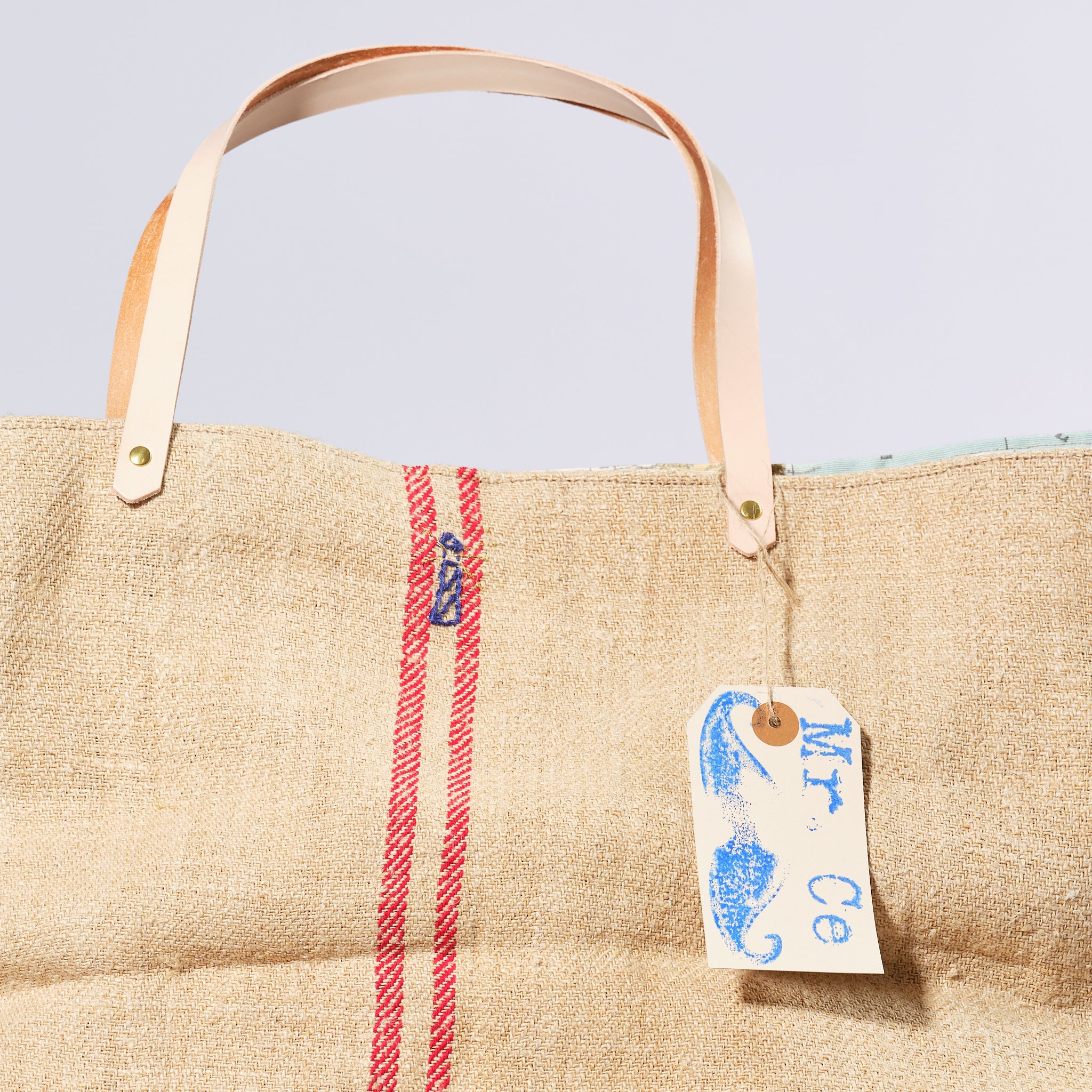 Beige tote bag with a red stripe and a visible brand tag on a light blue background