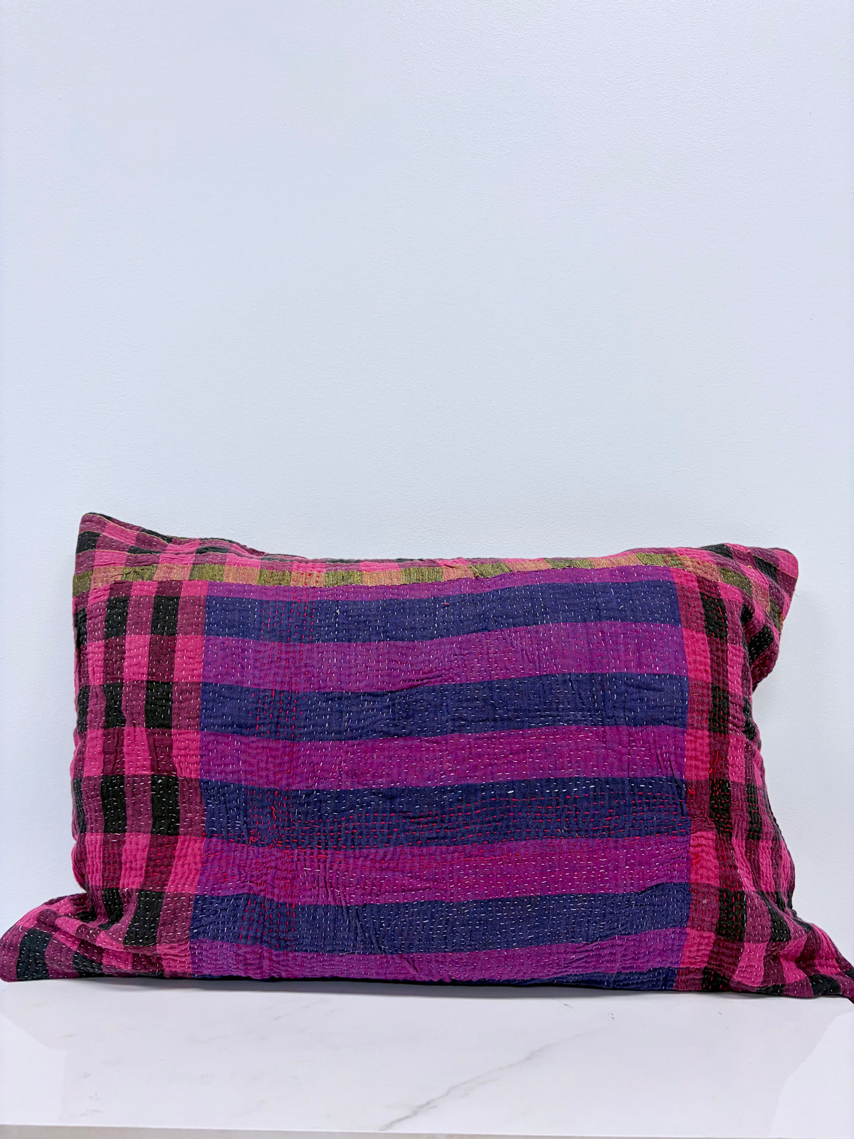 A rectangular throw pillow with a vibrant magenta pink and dark blue plaid pattern, accented with thin black and green stripes, resting on a white marble surface against a light gray wall.