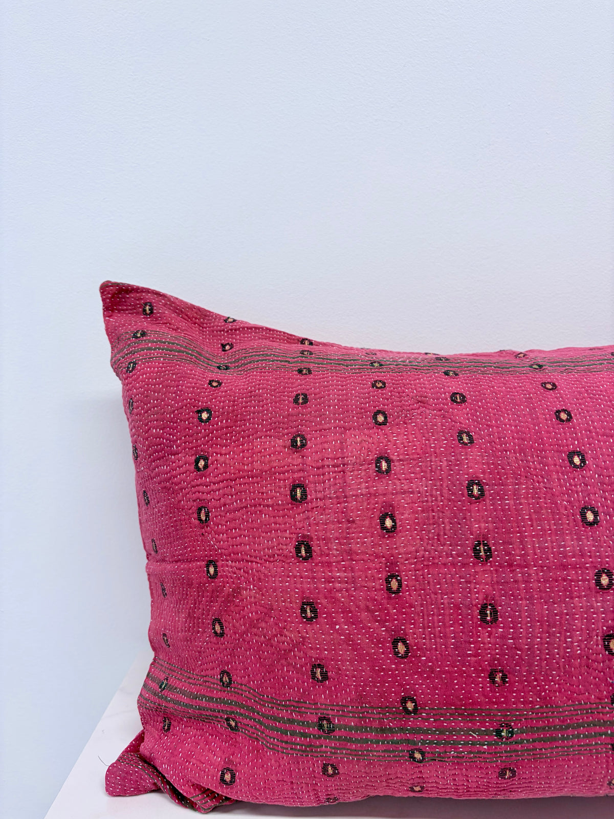 A close-up of a textured pink throw pillow with black embroidered geometric patterns and stripes, resting on a white surface against a light blue-gray background.