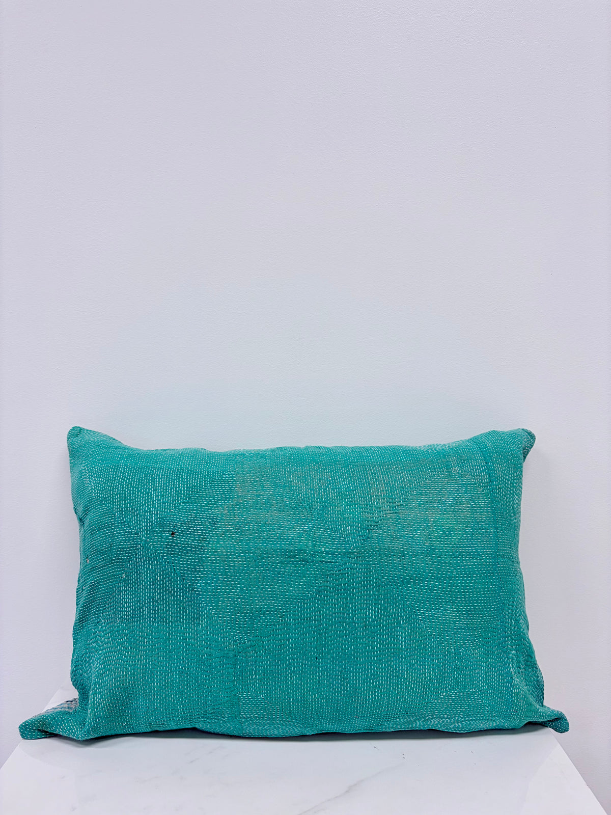A square teal green textured throw pillow made of a linen-like fabric, resting on a white marble surface against a light gray wall.