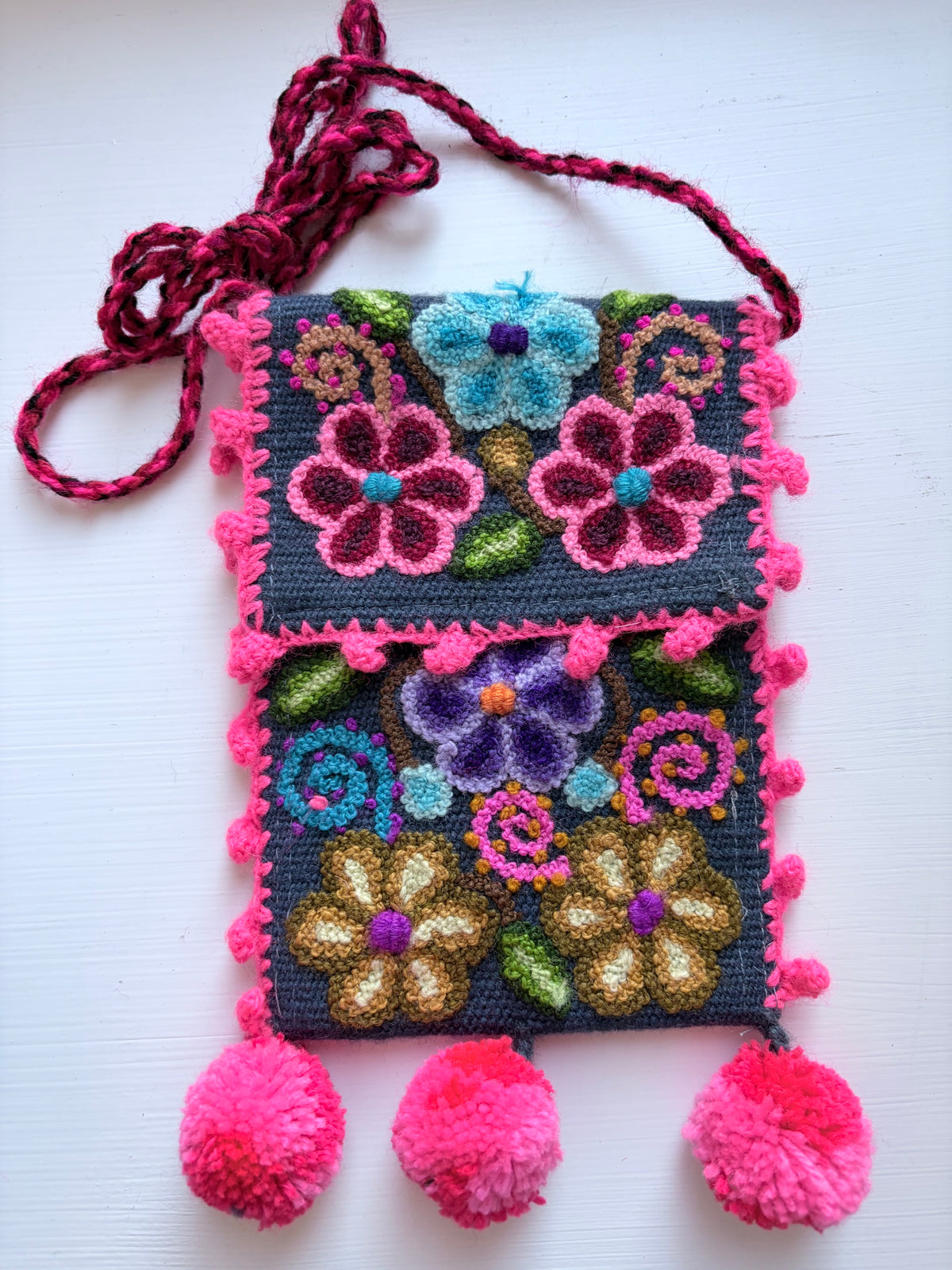 A handmade embroidered crossbody bag with a dark gray woven base, vibrant multicolor floral embroidery, hot pink pom-pom trim, and a matching pink braided strap, displayed on a white background.