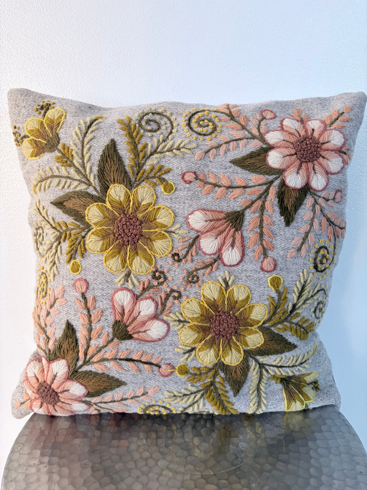 A square decorative throw pillow with a textured gray fabric base and colorful floral embroidery in yellow, pink, and olive green, resting on a dark reflective surface against a white wall.