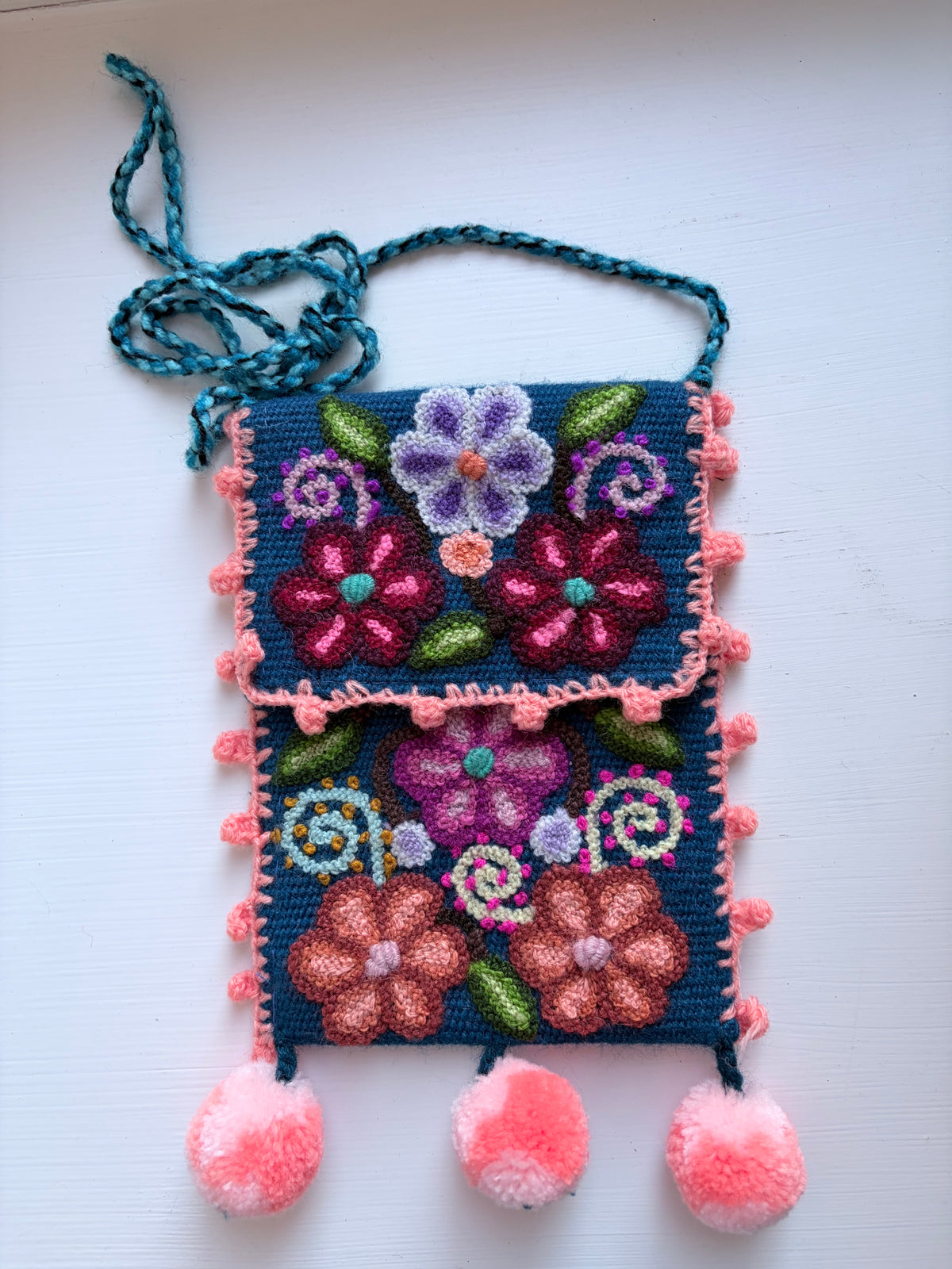 A handmade embroidered pouch with a dark blue base, featuring colorful floral and swirl patterns in pink, purple, green, and orange. The pouch has a teal braided strap, pink pom-pom trim, and three fluffy pink pom-poms hanging from the bottom, displayed on a white background.