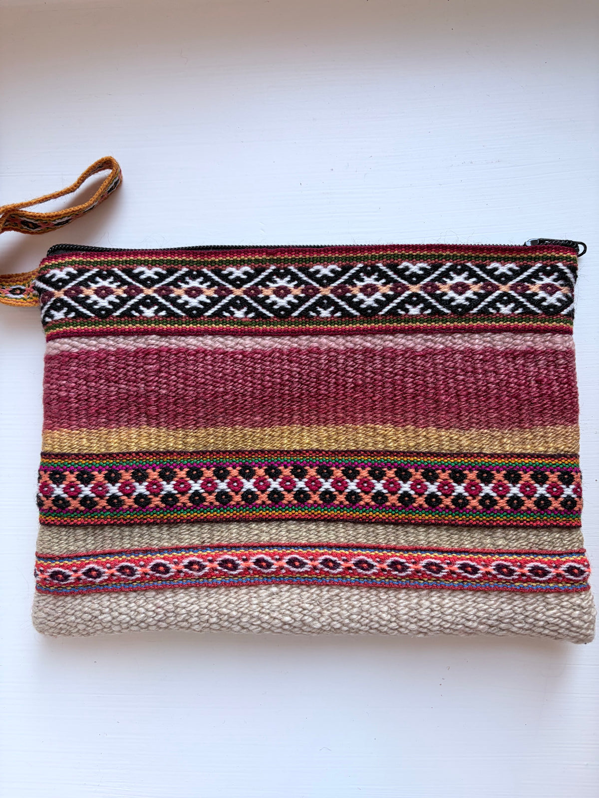 A woven zippered pouch with a multicolor Southwestern geometric pattern in red, black, white, and beige, featuring a brown woven wrist strap, laid flat on a white background.