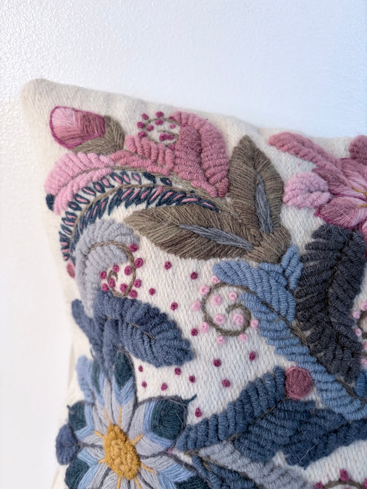 Close-up of a decorative embroidered throw pillow featuring a floral and leaf pattern in dusty pink, blue, gray, and yellow on a cream background.
