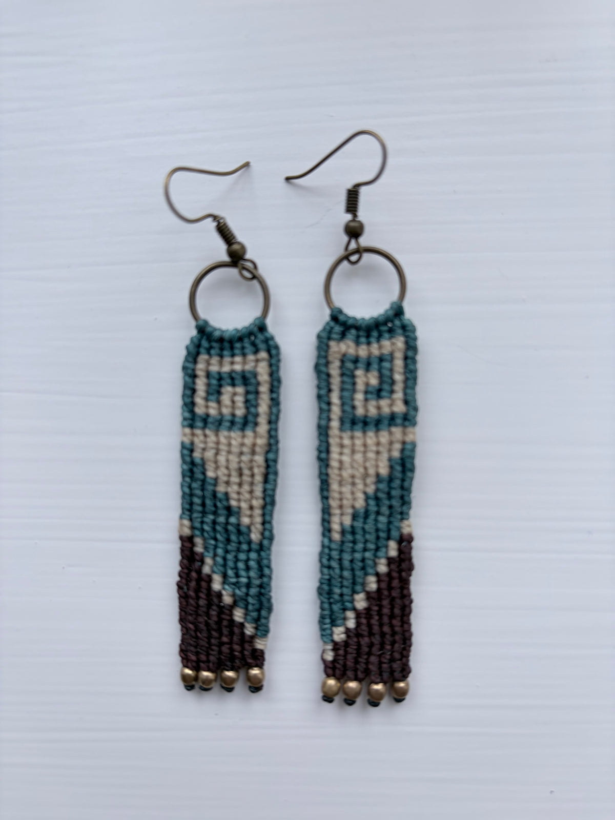 A pair of handmade beaded dangle earrings with geometric patterns in teal, cream, and dark brown, hanging from dark metal fishhook wires on a white background.