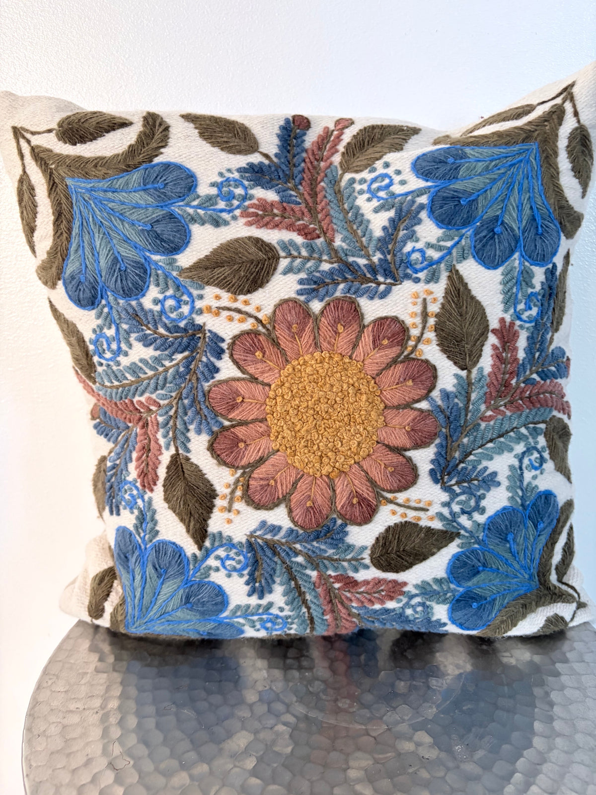 A square decorative throw pillow with a detailed floral and botanical embroidery pattern in blue, brown, rust, and gold on a white background, resting on a textured metallic surface.