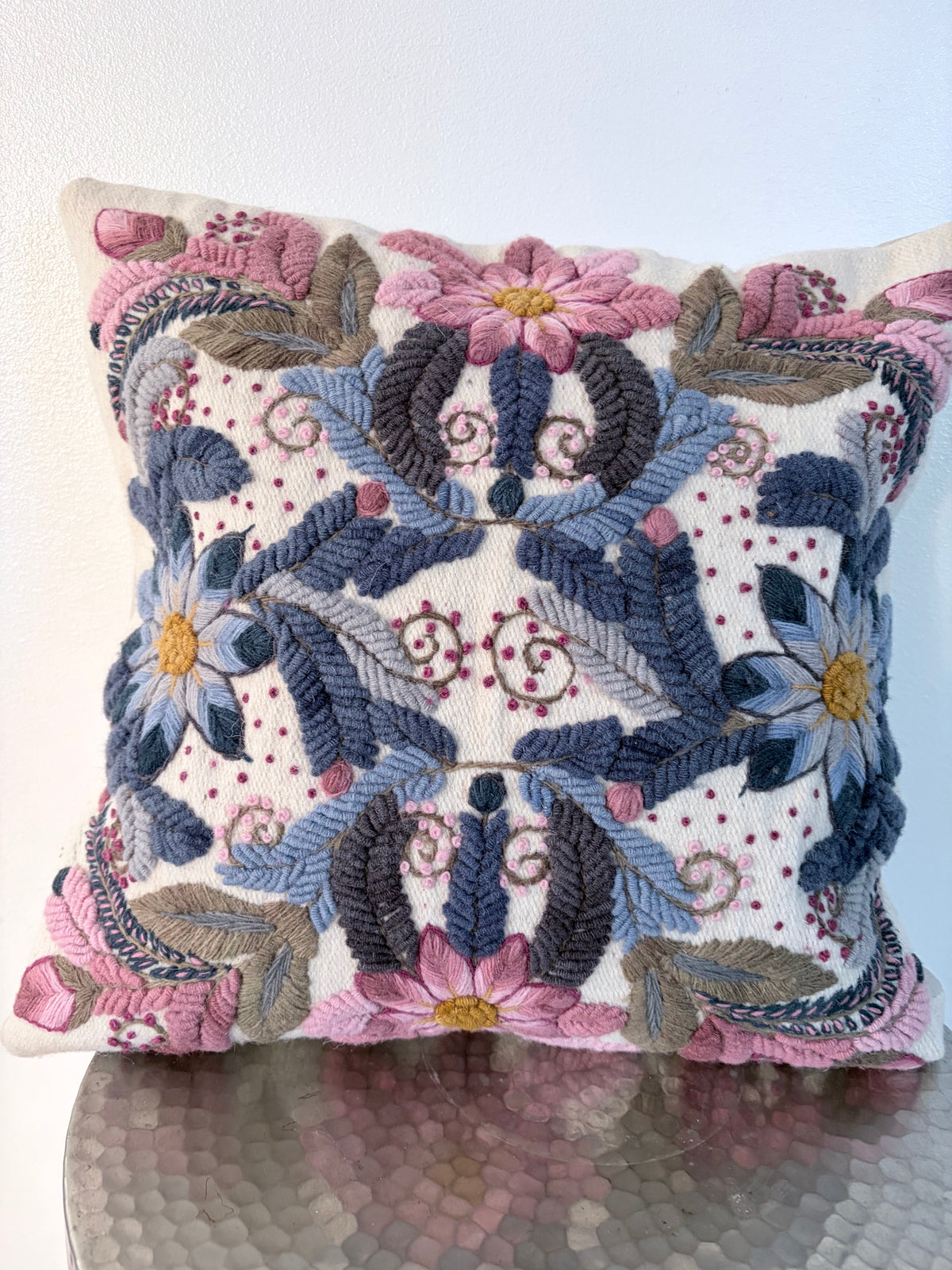 An ornate square throw pillow featuring intricate floral and scroll embroidery in shades of blue, pink, gray, and gold on a cream background, resting on a reflective metallic surface.