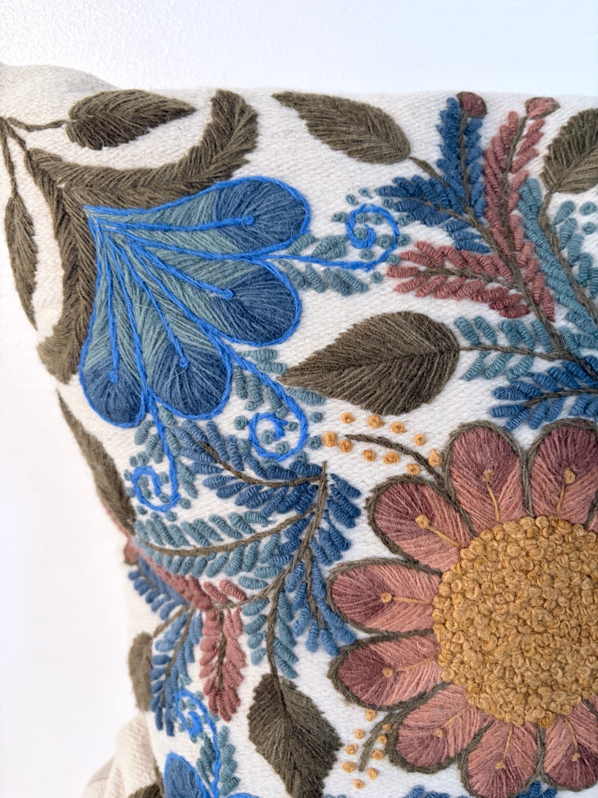 Close-up of a decorative embroidered throw pillow featuring a floral and leaf pattern in blue, dusty pink, olive green, and mustard yellow on a cream background.