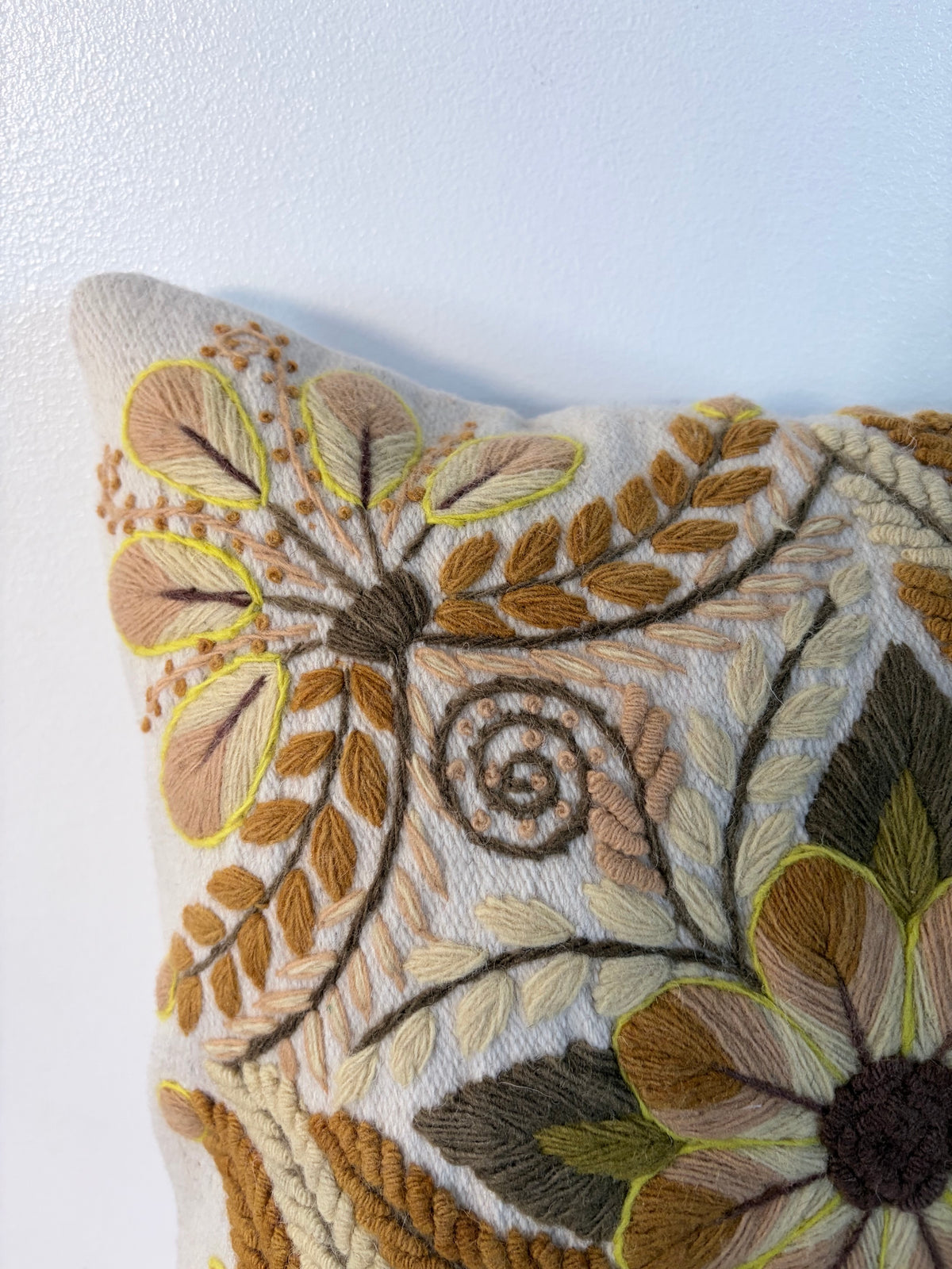 Close-up of a decorative throw pillow featuring intricate hand-embroidered floral and leaf patterns in earthy tones of brown, beige, olive green, and mustard yellow on a cream fabric background.