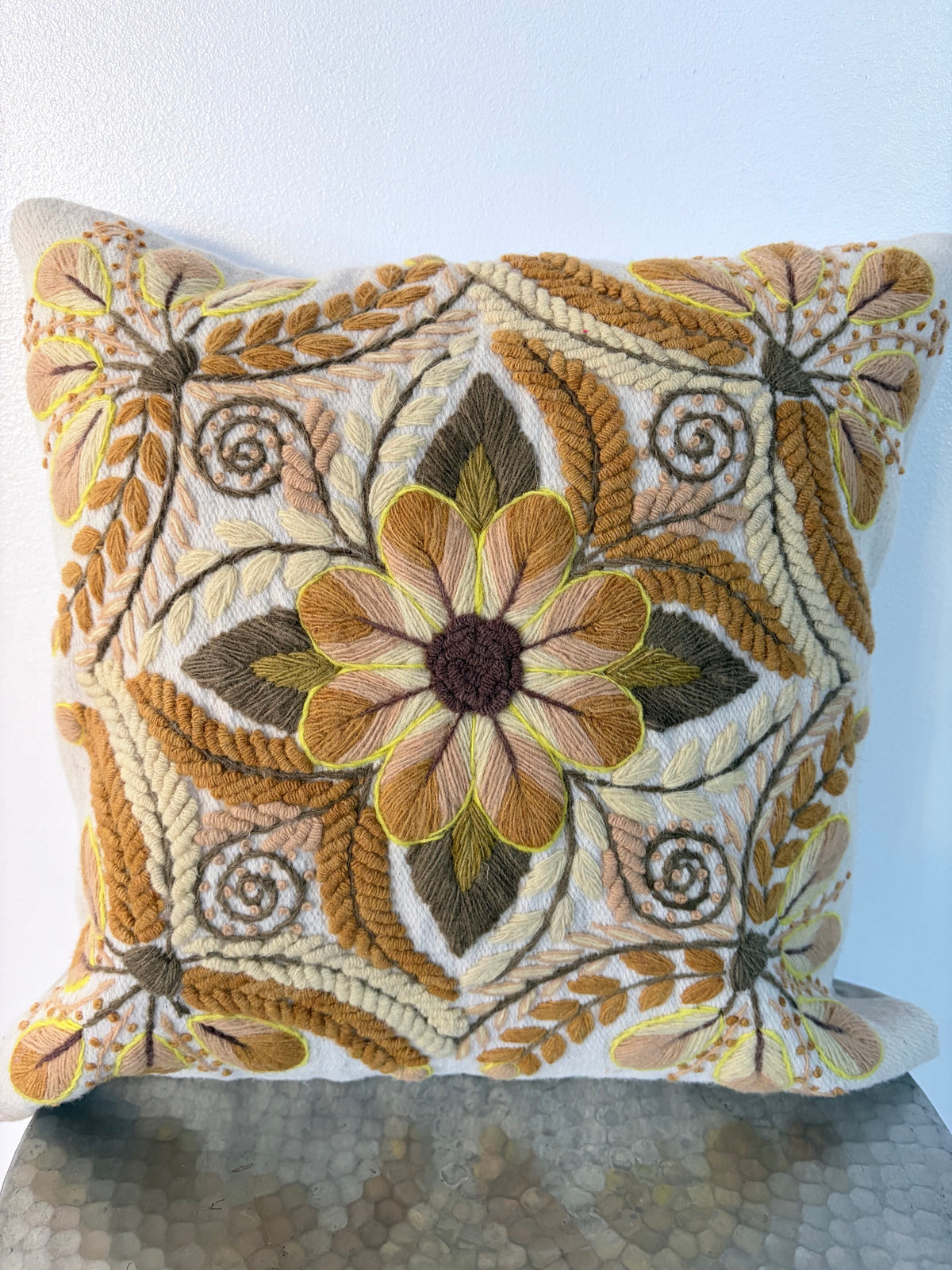 A square decorative throw pillow featuring intricate hand-embroidered floral and leaf patterns in mustard yellow, olive green, cream, and brown, set against a plain white wall.