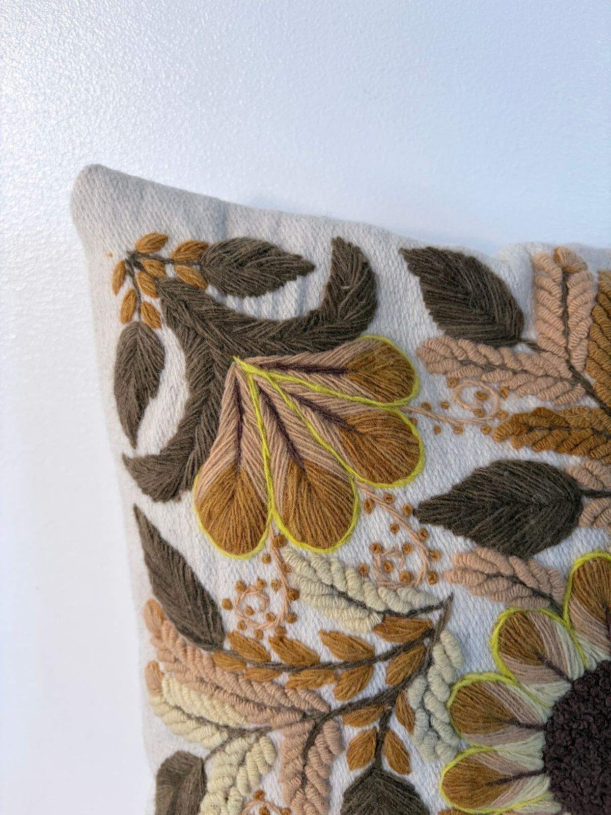A close-up of a decorative throw pillow featuring intricate hand-embroidered floral and leaf patterns in earthy tones of brown, mustard, beige, and olive green on a light cream fabric background.