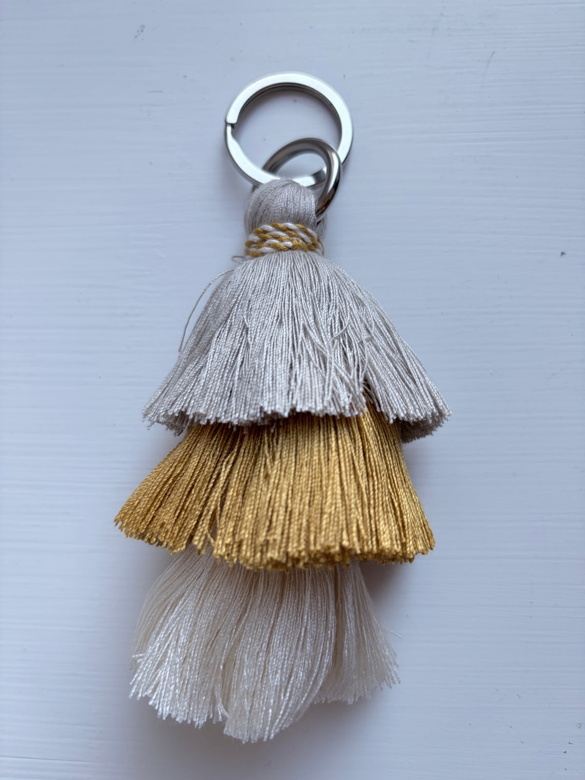 A three-tiered tassel keychain with a silver split ring, featuring layers of light gray, metallic gold, and off-white threads on a light gray background.