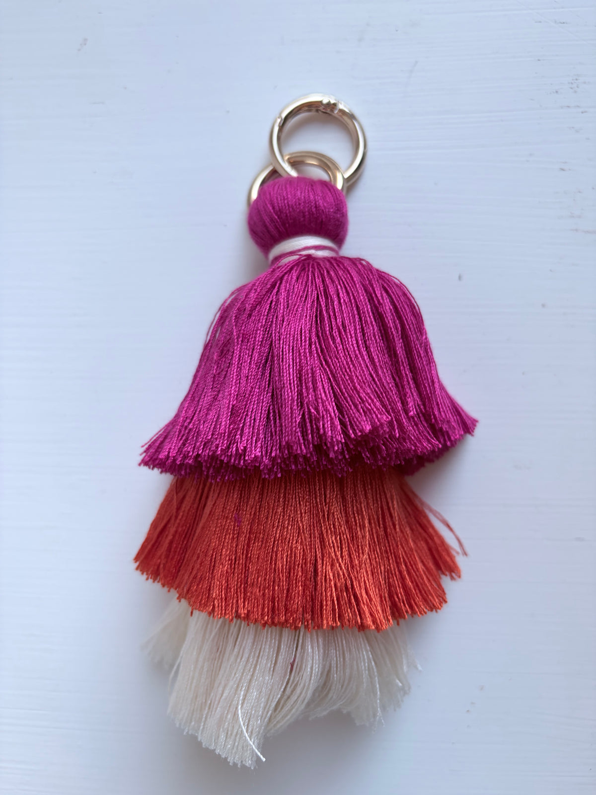 A decorative tassel keychain with three tiers of fringed threads in magenta pink, orange, and off-white, attached to gold metal rings, shown on a light blue background.