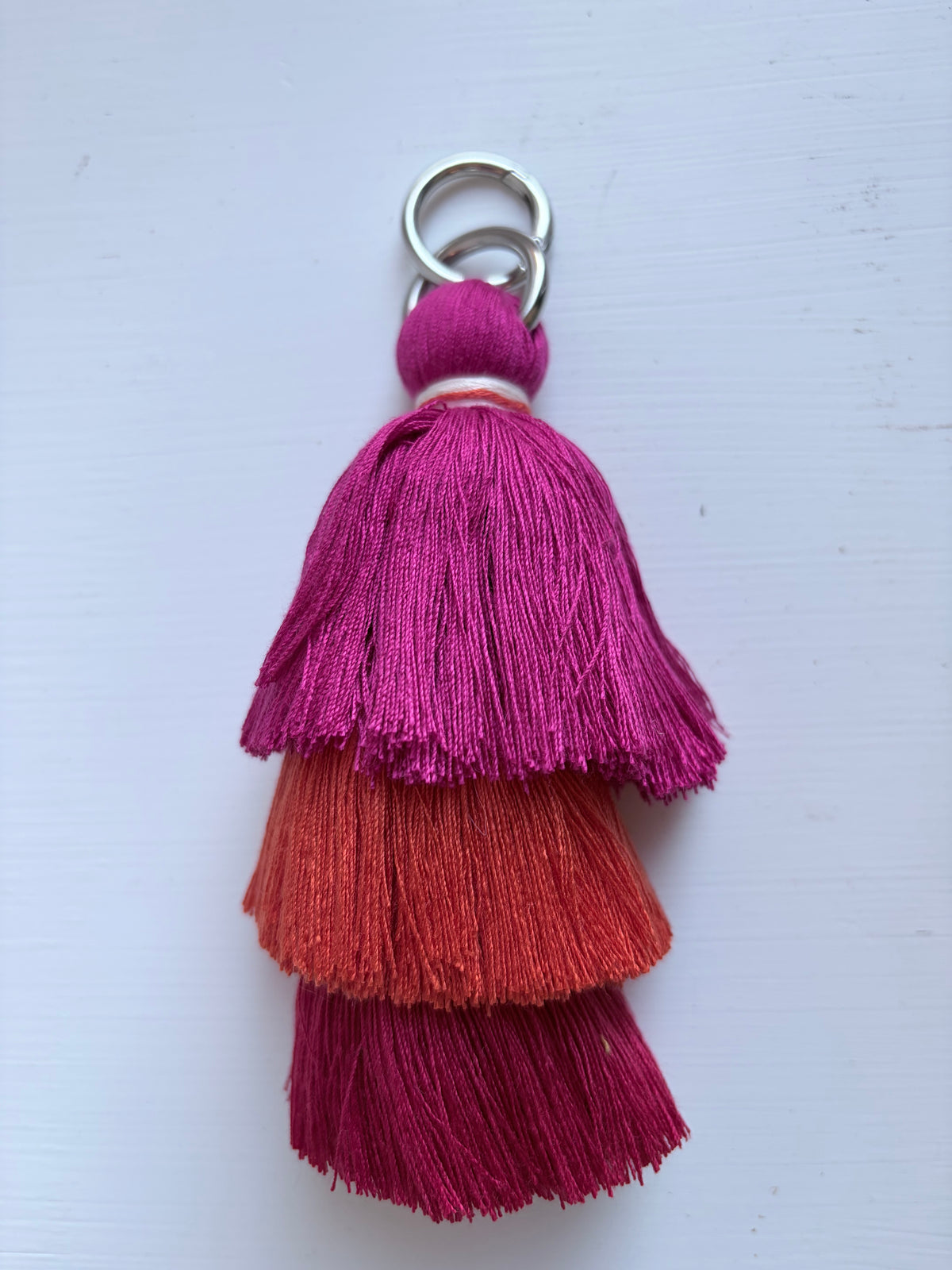A three-tiered tassel keychain with vibrant magenta, orange, and deep pink tassels, attached to a silver metal key ring, set against a light off-white background.