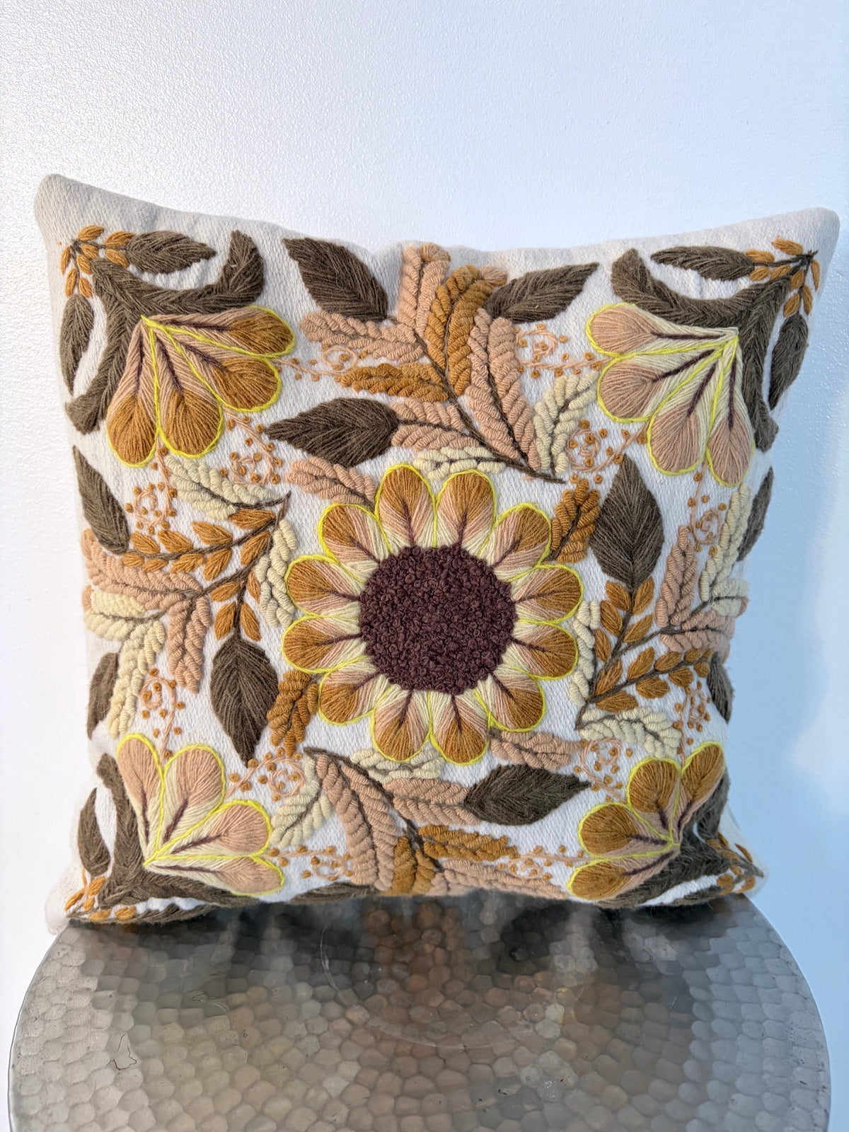 A square decorative throw pillow with an intricate embroidered floral and leaf pattern in earthy tones of brown, beige, mustard yellow, and dark plum, resting on a reflective metallic surface against a light blue wall.
