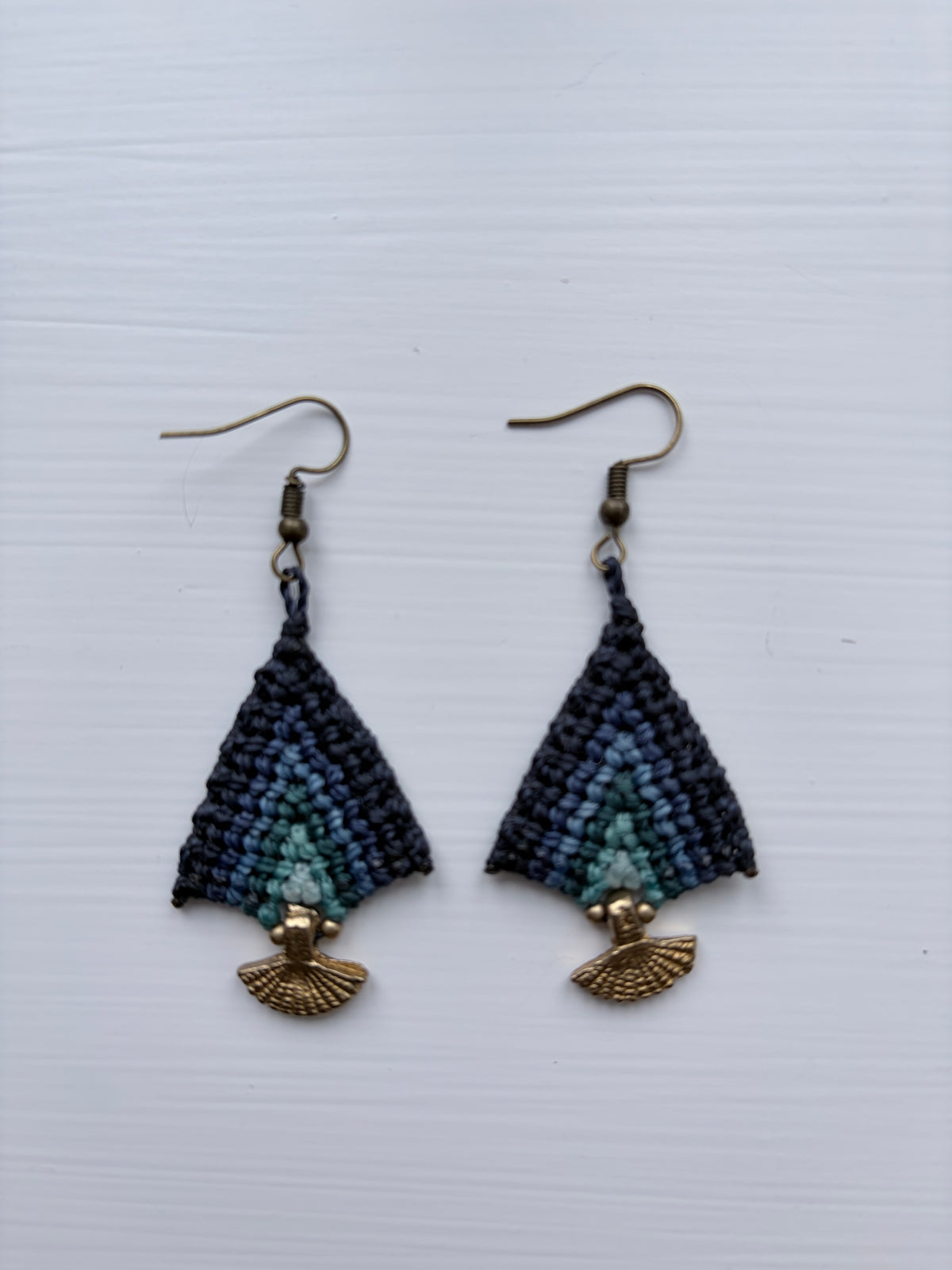 A pair of handmade beaded dangle earrings with a dark blue and teal geometric pattern, finished with gold-tone fishhook wires and fan-shaped charms, displayed on a light gray textured background.