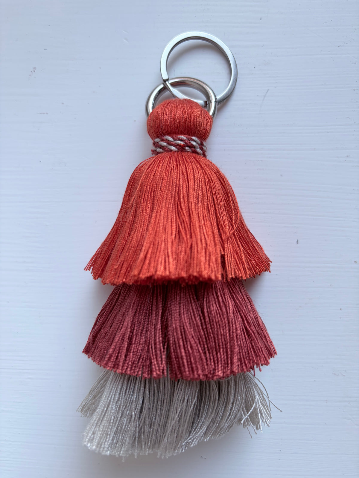 A decorative tassel keychain with three tiers of fringed thread in rust orange, dusty rose, and light gray, attached to silver metal key rings on a white background.
