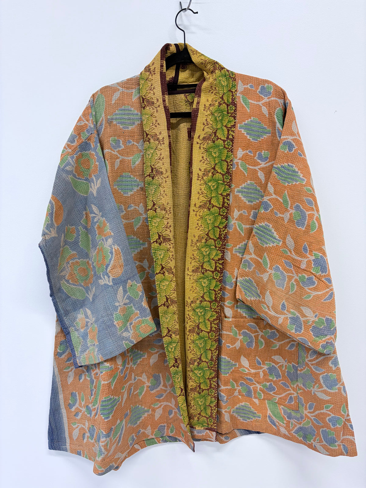 A lightweight open-front kimono-style jacket with a mustard yellow base, featuring green and blue floral patterns, hanging on a black hanger against a plain white wall.