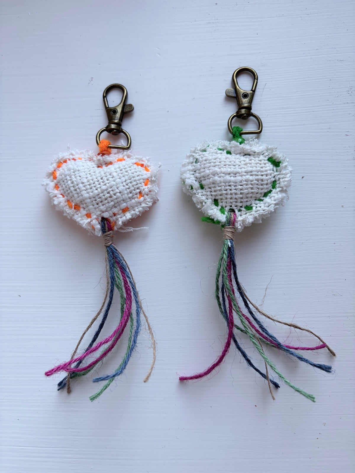 Two handmade embroidered heart keychains on a white background. Each keychain features a white linen-textured heart with one colored border (orange on the left, green on the right), a brass clasp, and a long multicolored tassel.