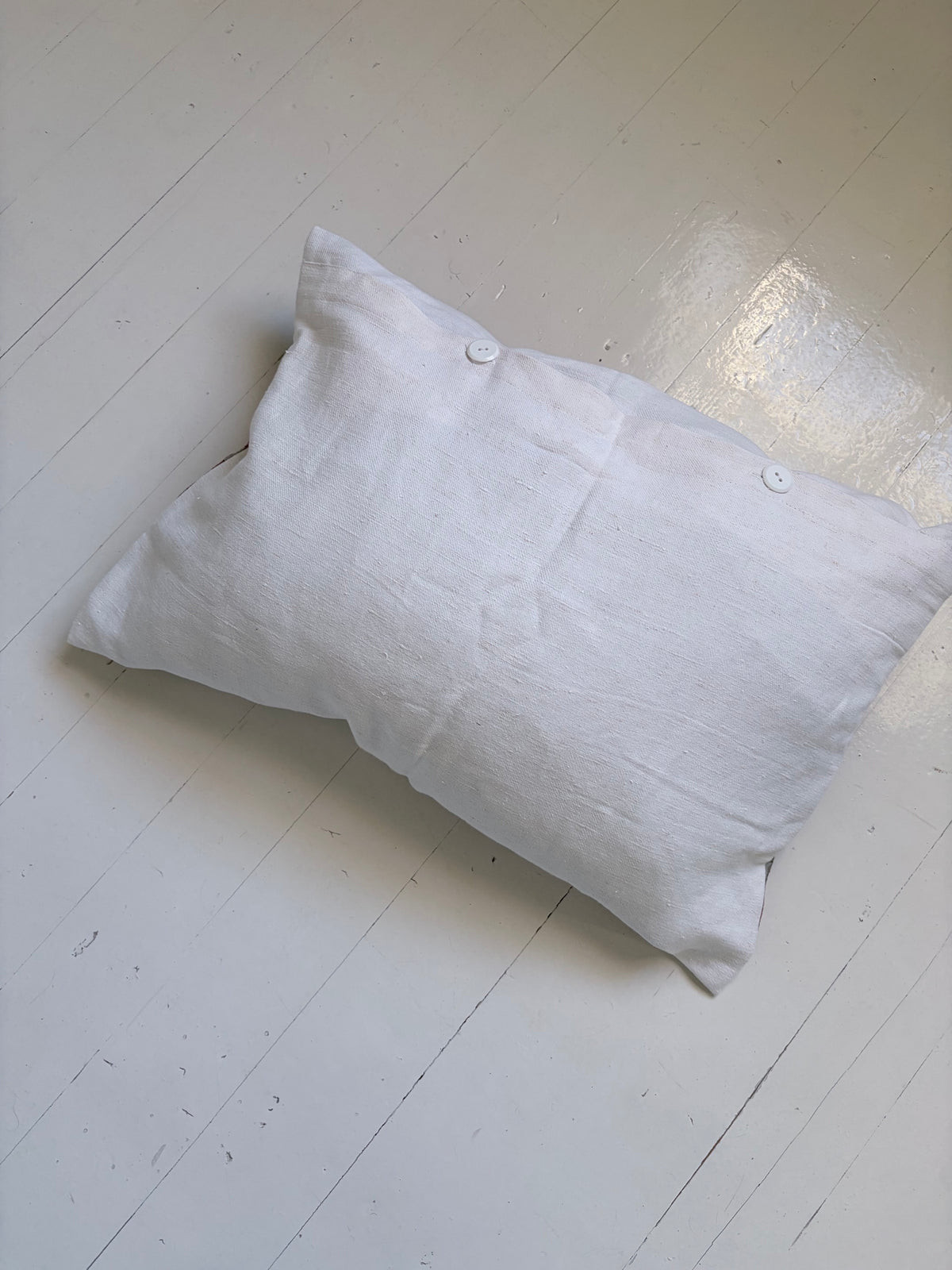 A white linen pillow with two decorative buttons on the front, resting on a white wooden floor.