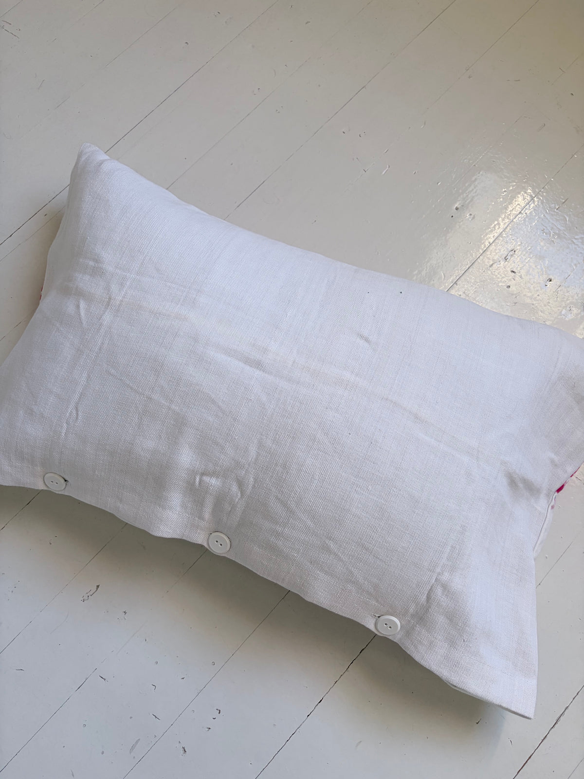 A white linen pillow sham with three decorative white buttons along the bottom edge, laid flat on a white wooden floor.