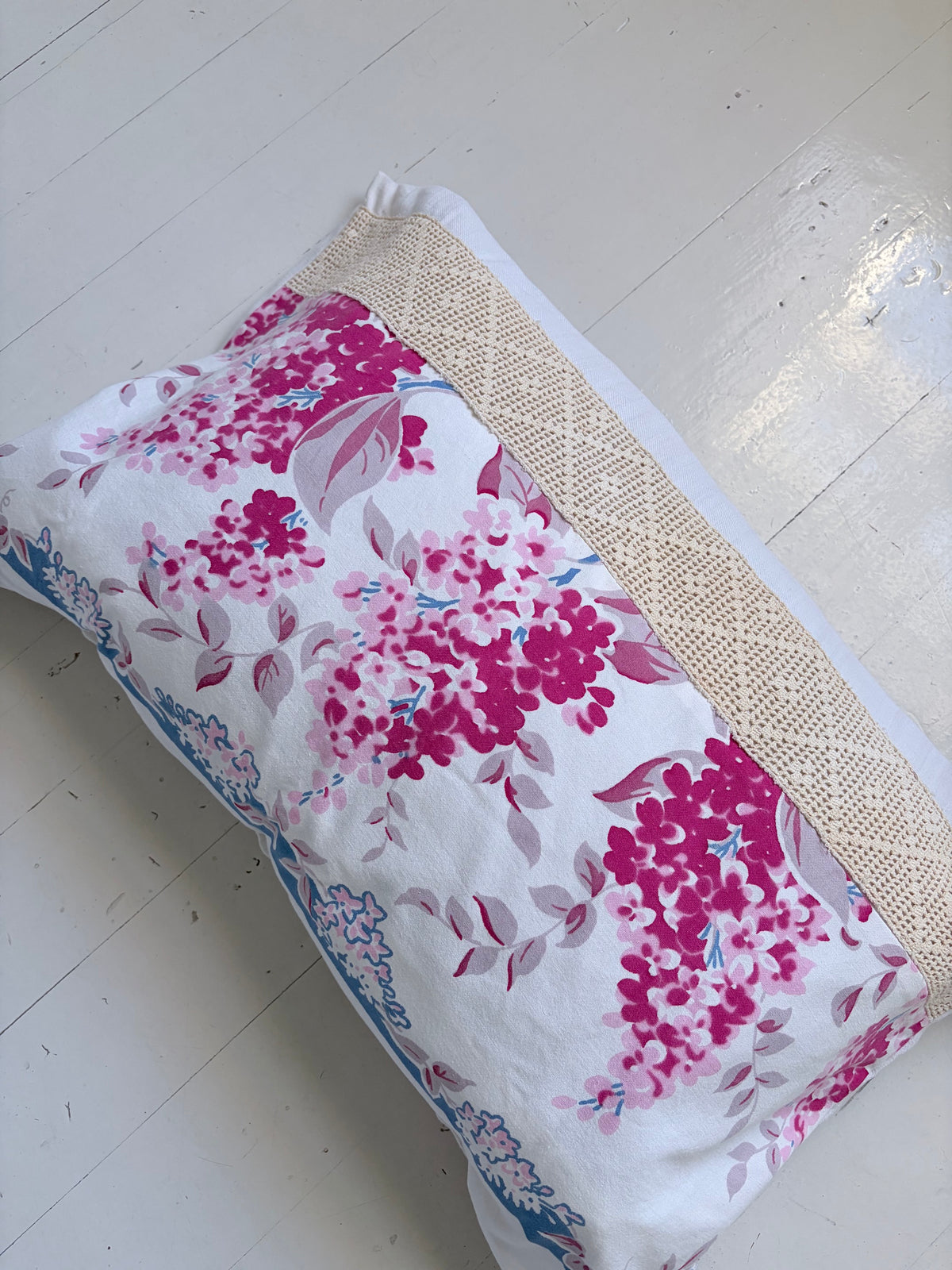 A rectangular pillow with a white background, featuring a vibrant pink and magenta floral print on one side and a beige woven textured band on the other, resting on a white wooden floor.