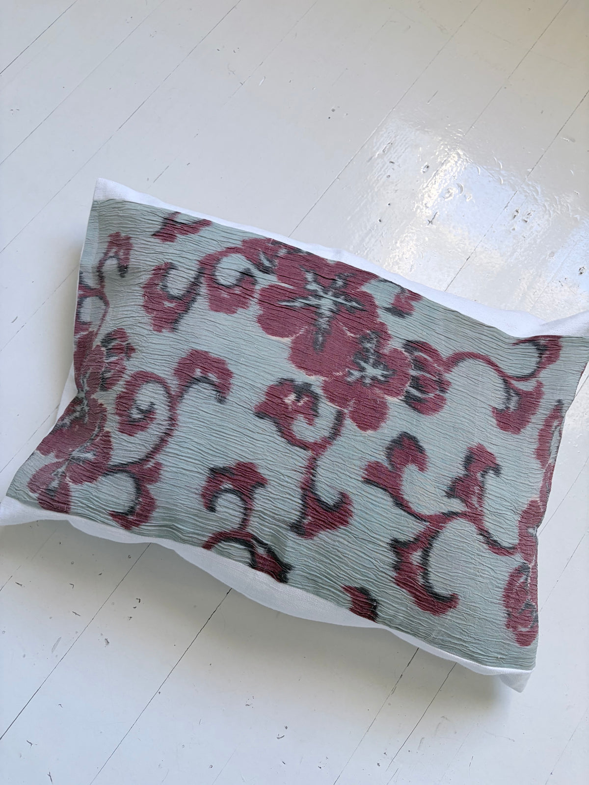 A rectangular decorative pillow with a textured, crinkled fabric cover featuring a burgundy floral and scroll pattern on a muted sage green background, resting on a white wooden floor.