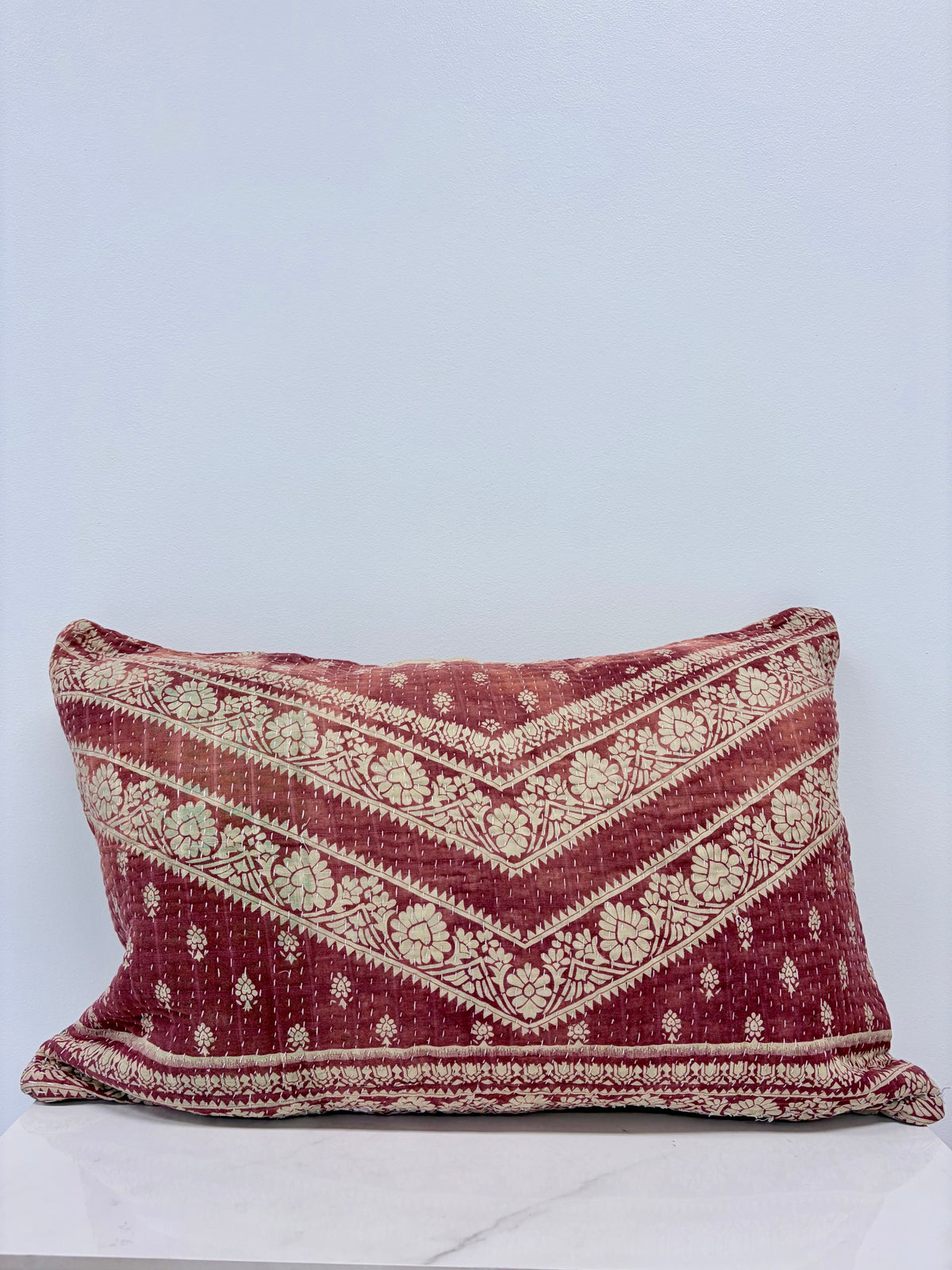A square throw pillow with a distressed, vintage-style bohemian pattern in deep red and cream, resting on a white marble surface against a light gray background.