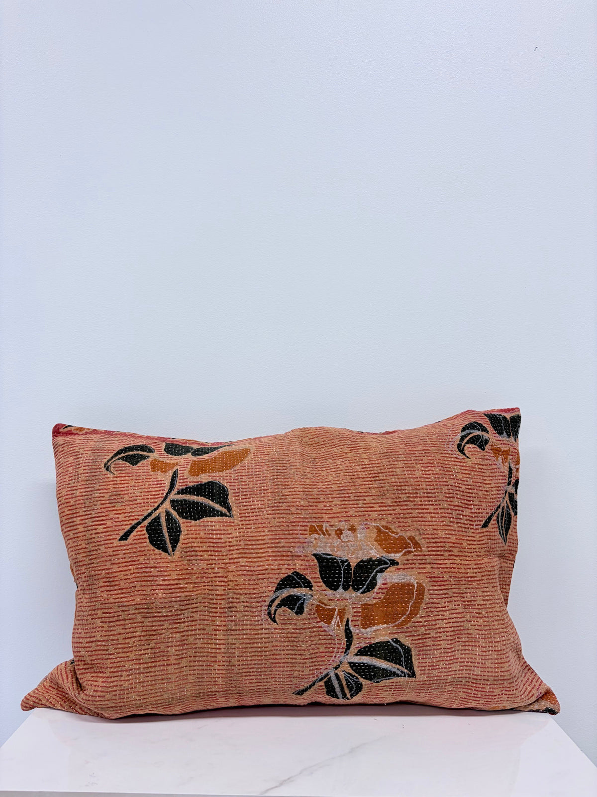 A square decorative throw pillow with a textured terracotta orange fabric featuring a black and mustard yellow floral pattern, resting on a white marble surface against a light blue-gray background.